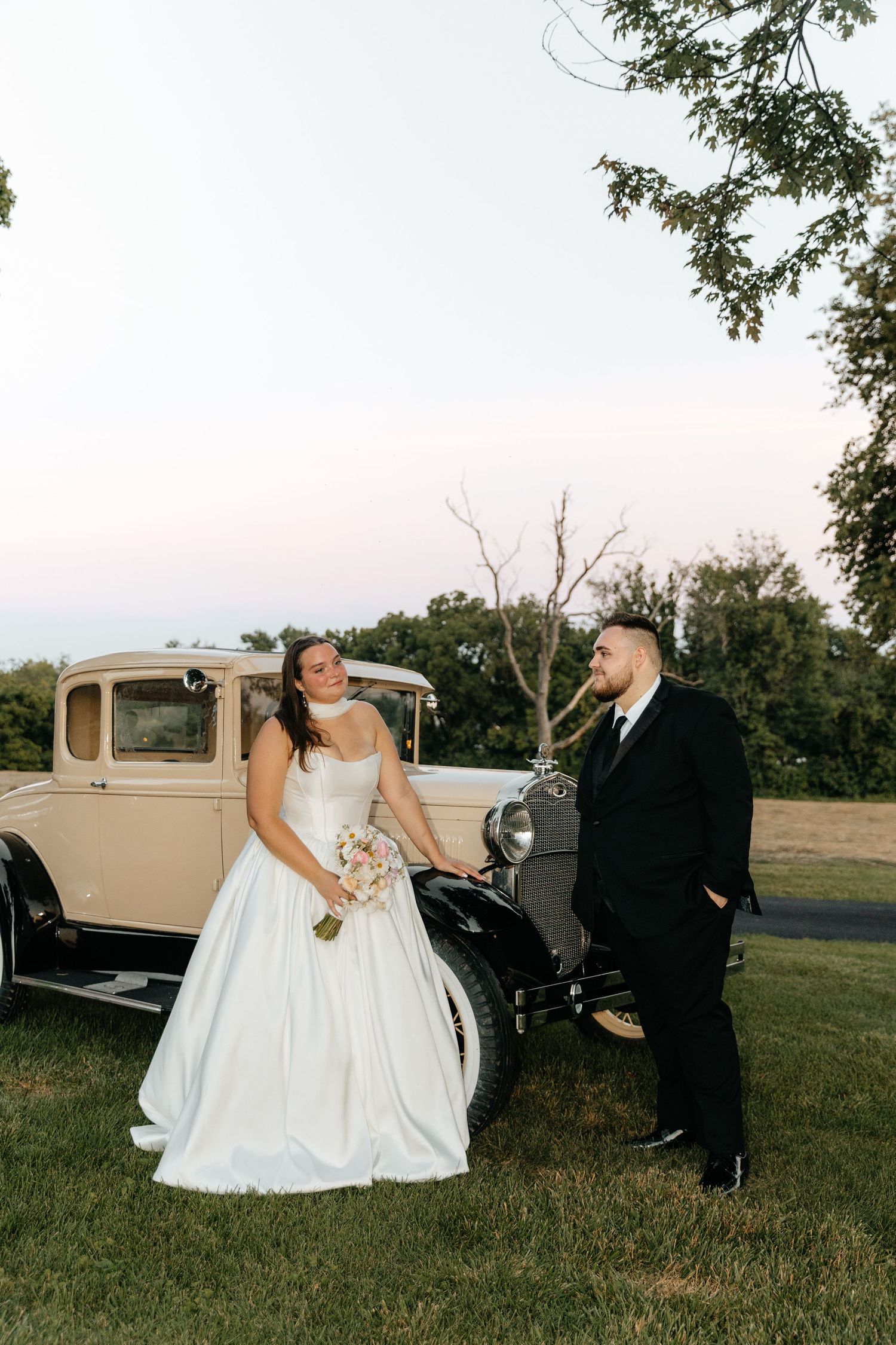 Wedding couple shares joyful moment beside vintage cream car during outdoor photoshoot.