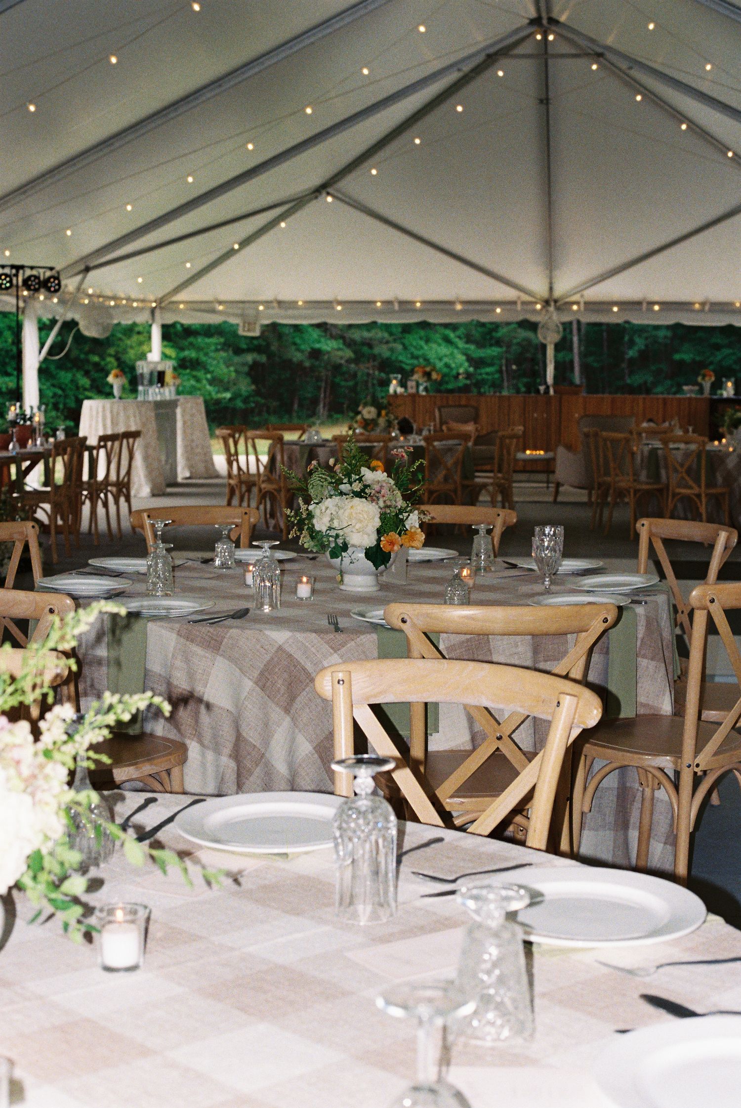 Elegant outdoor wedding reception under a white tent with string lights, wooden cross-back chairs, and floral centerpieces on round tables.
