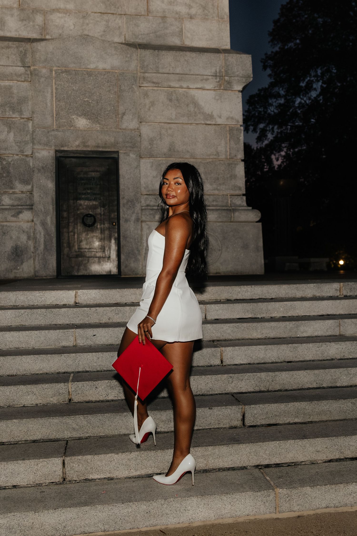 Woman in white mini dress holds red graduation cap on monument steps at night.