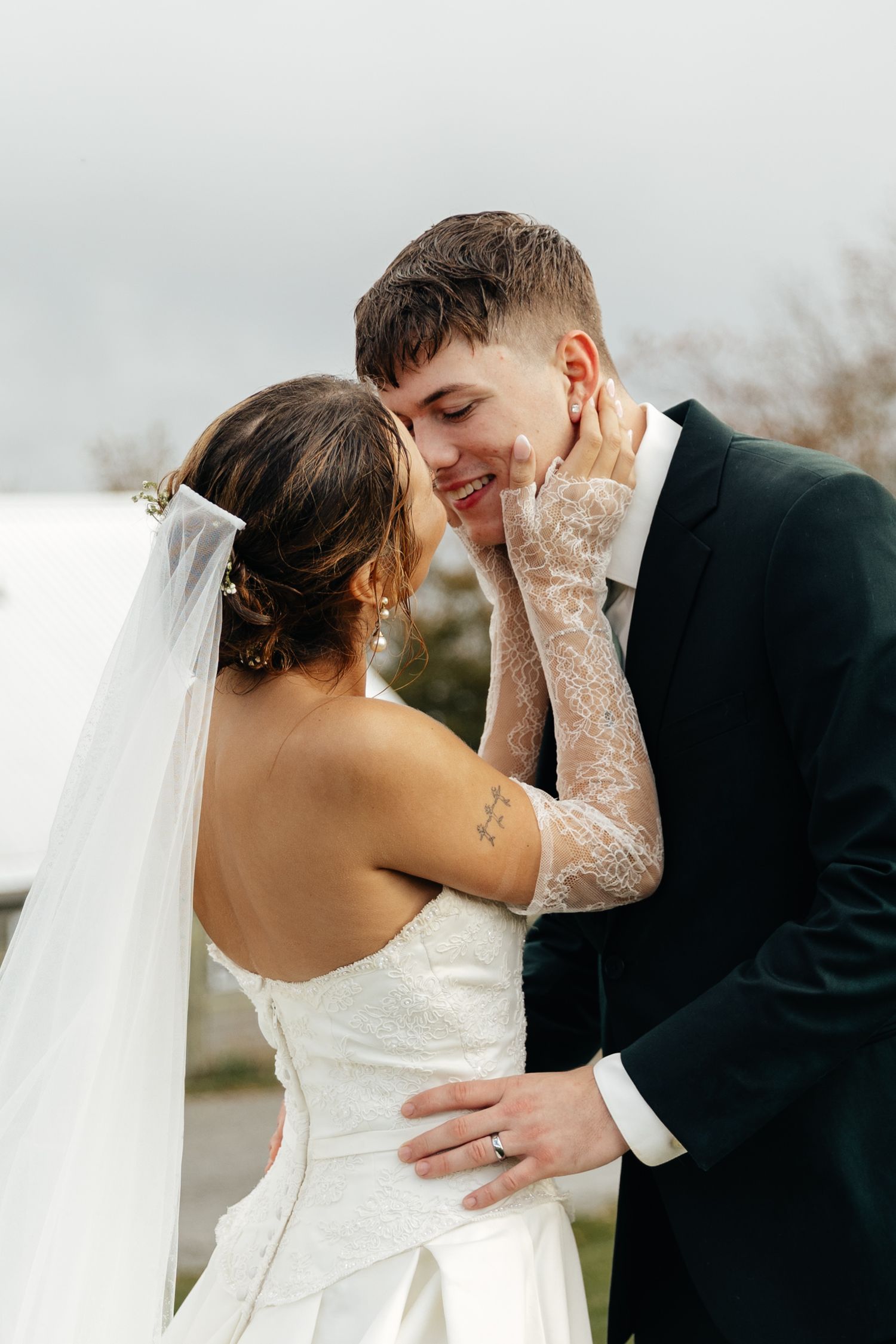 Bride in lace gloves tenderly holds groom's face as they share an intimate moment in wedding attire outdoors.