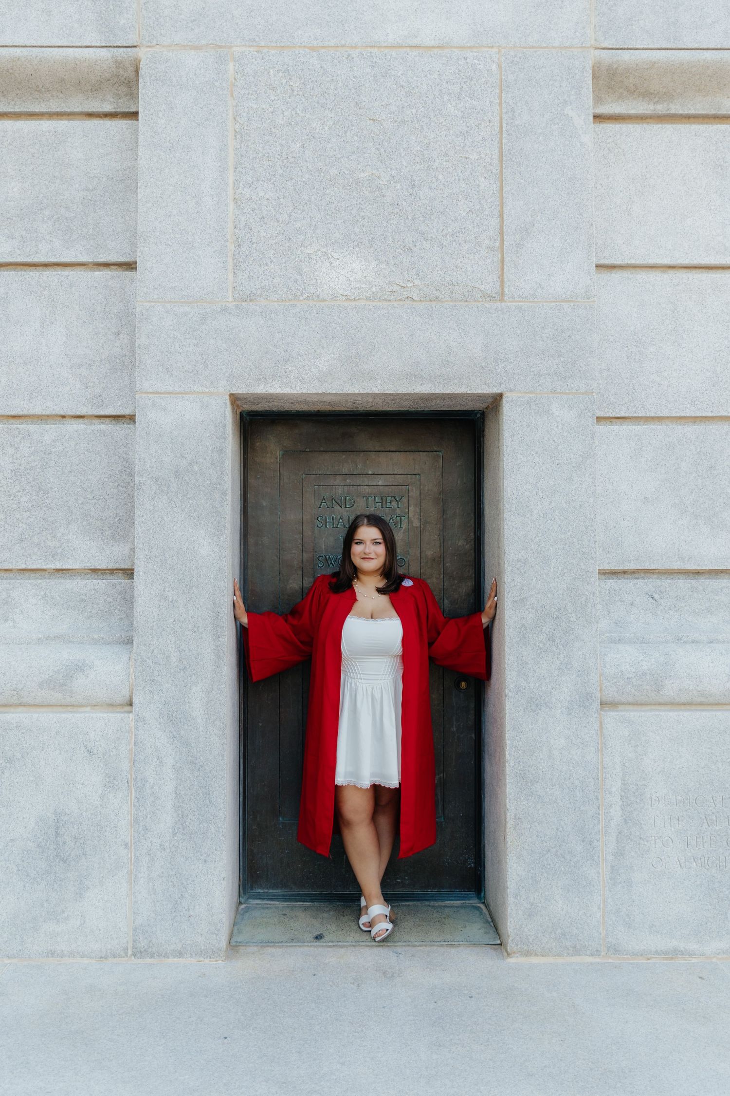 A young woman in a white dress and red graduation gown stands in a stone doorway, smiling at the camera.