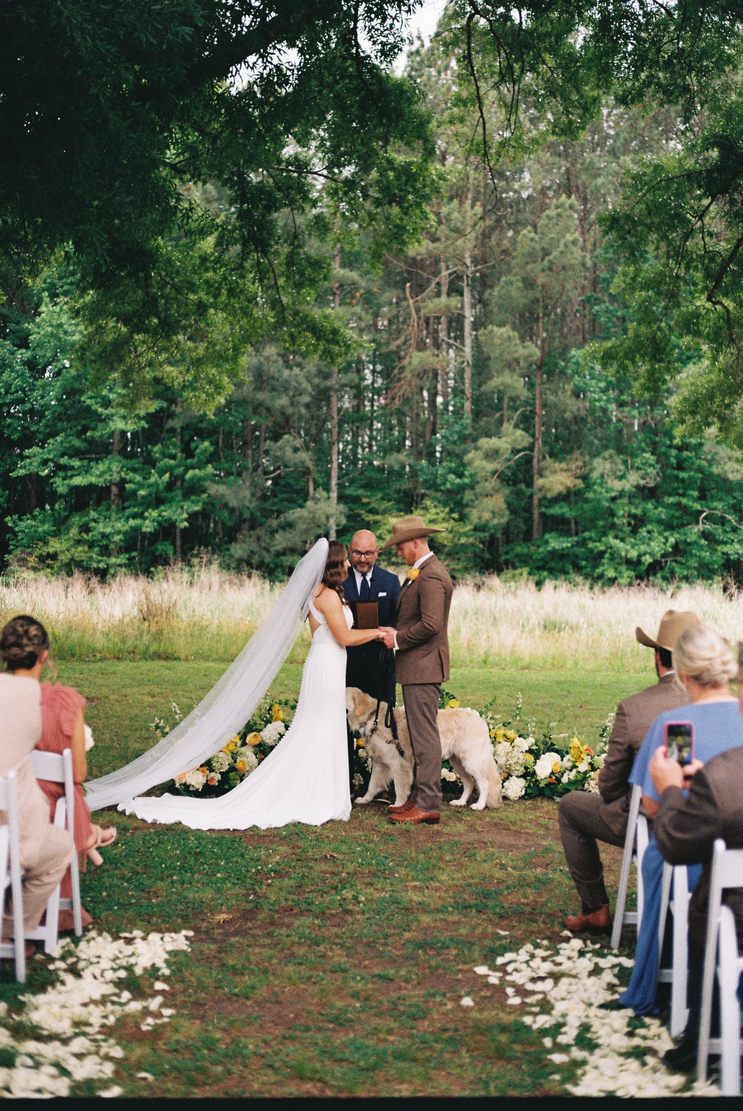 Outdoor wedding ceremony in lush forest setting with bride in white gown and veil, groom in brown suit, and golden dog at altar.