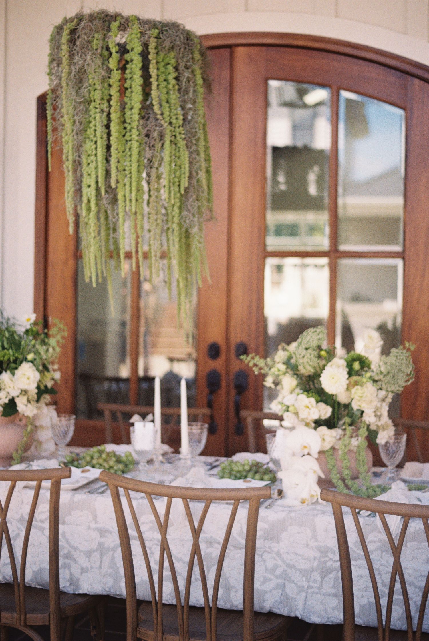 Elegant wedding reception table settings with white linens, floral centerpieces and hanging greenery in a rustic venue.