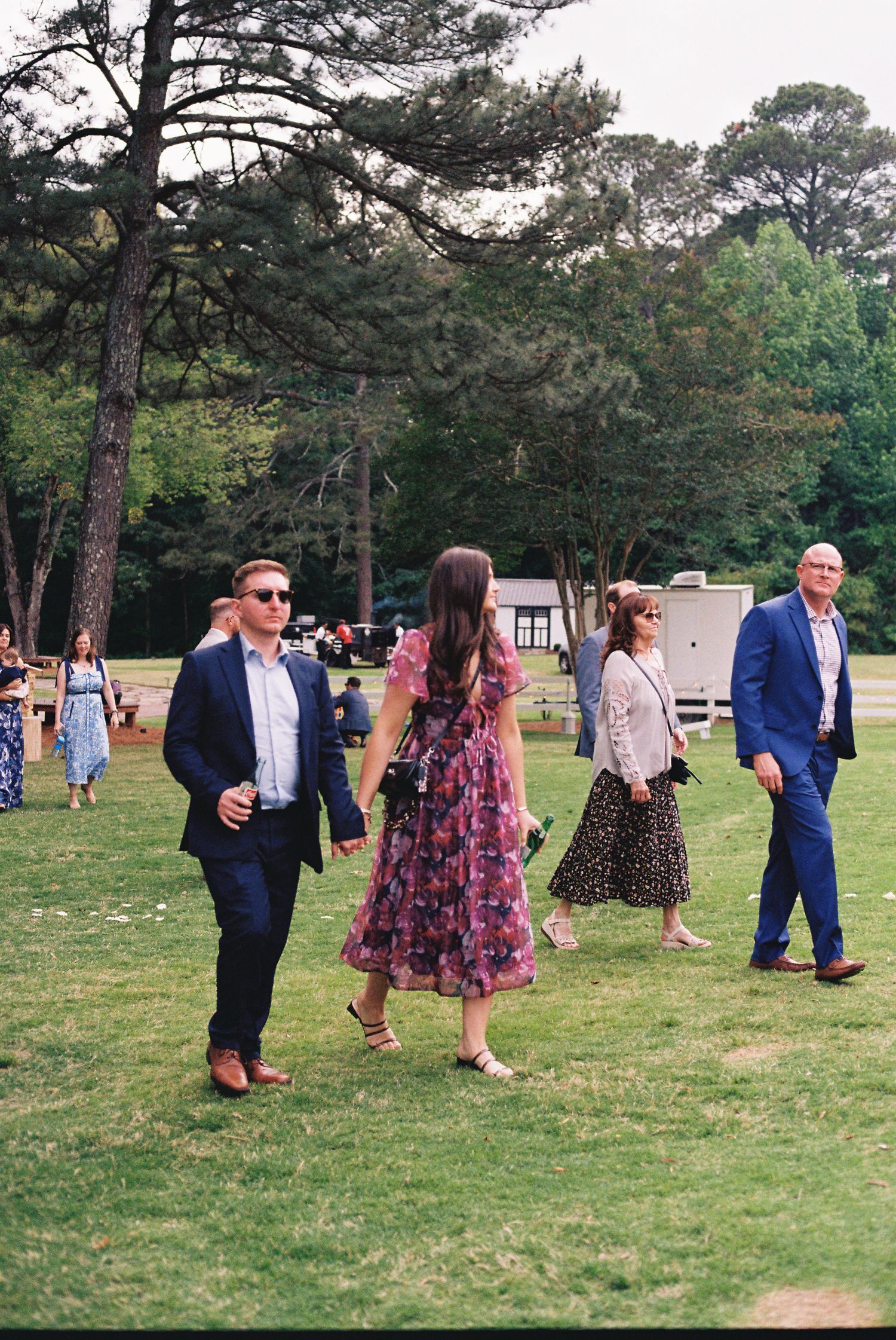 Wedding guests in formal attire walking across green lawn with tall pine trees in background.