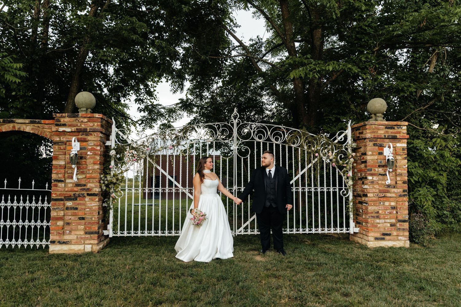 Wedding couple stands in front of ornate white iron gates and brick pillars.