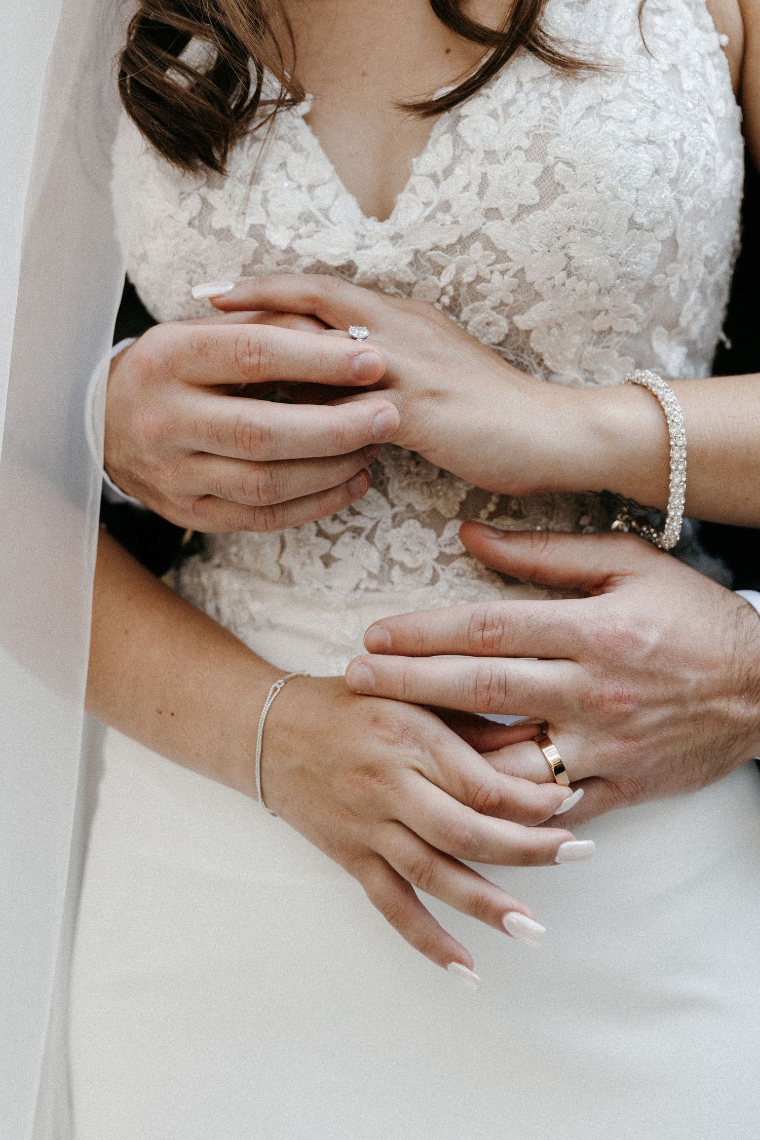 Close-up of hands with wedding rings and bracelet against a white lace wedding dress bodice.