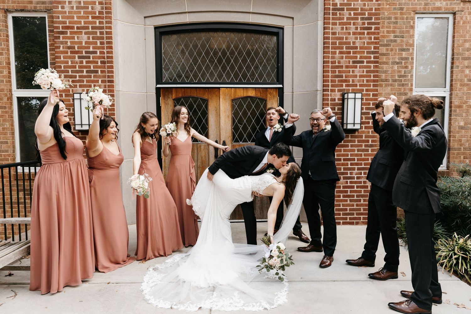 Wedding party lineup against brick building with bridesmaids in dusty rose and groomsmen in black.