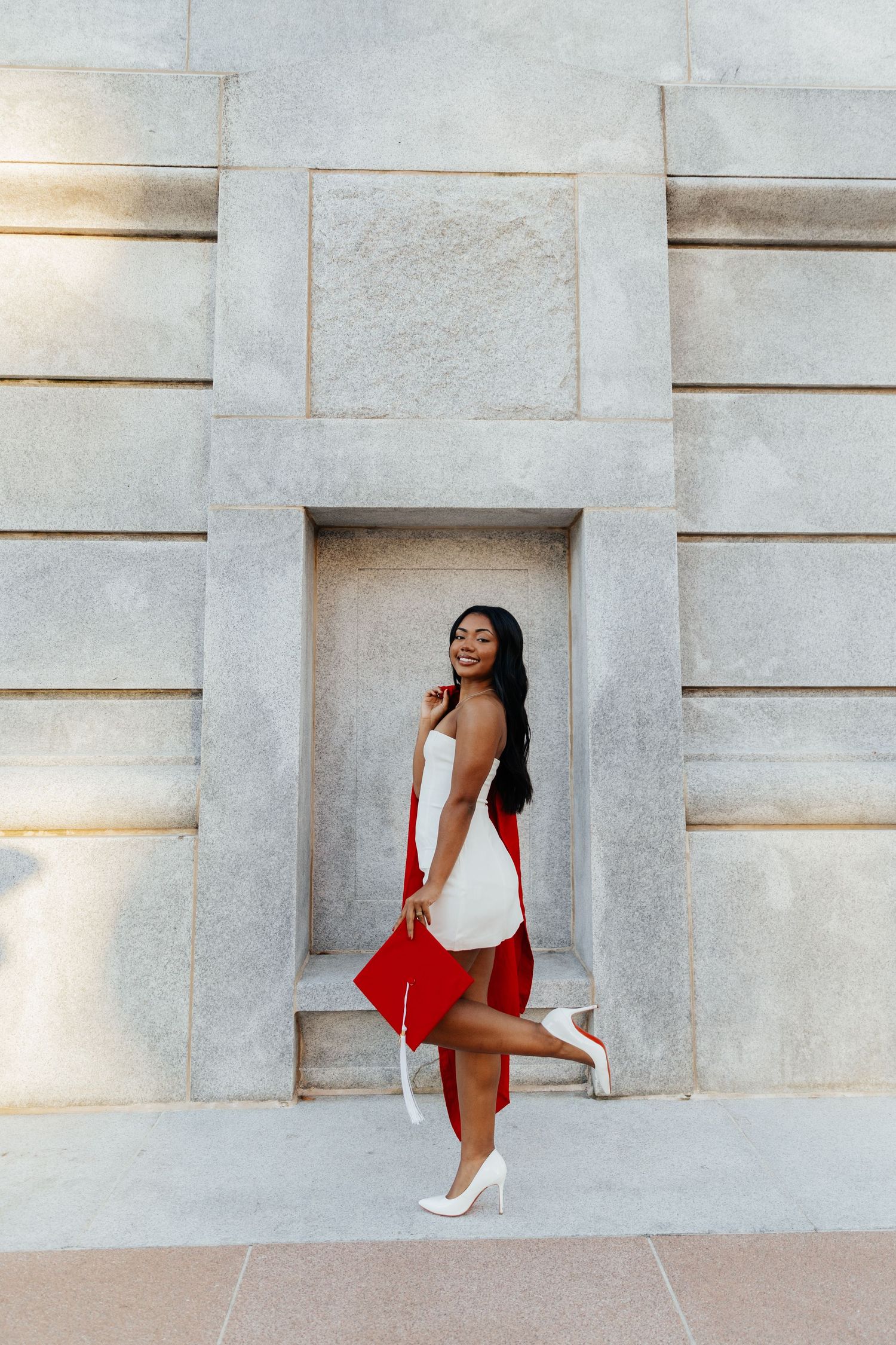 Young woman in white dress and red graduation gown poses confidently against stone wall holding cap.