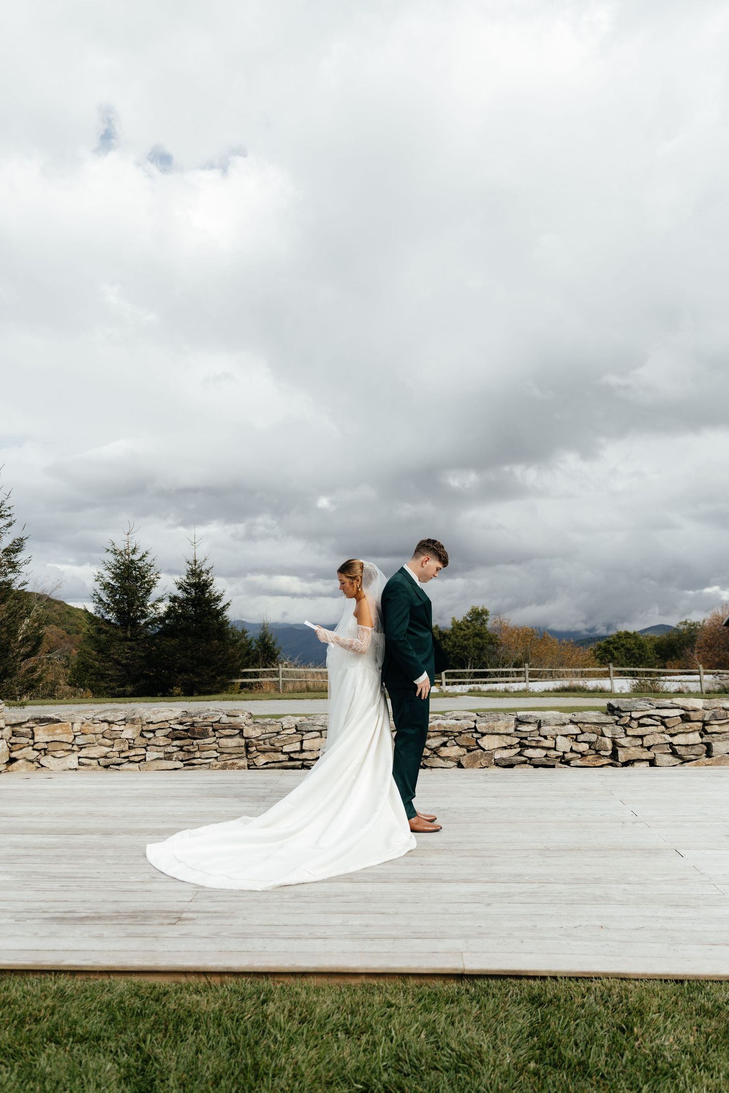Bride and groom share a tender moment on a wooden deck with a stone wall and mountain backdrop on their wedding day.