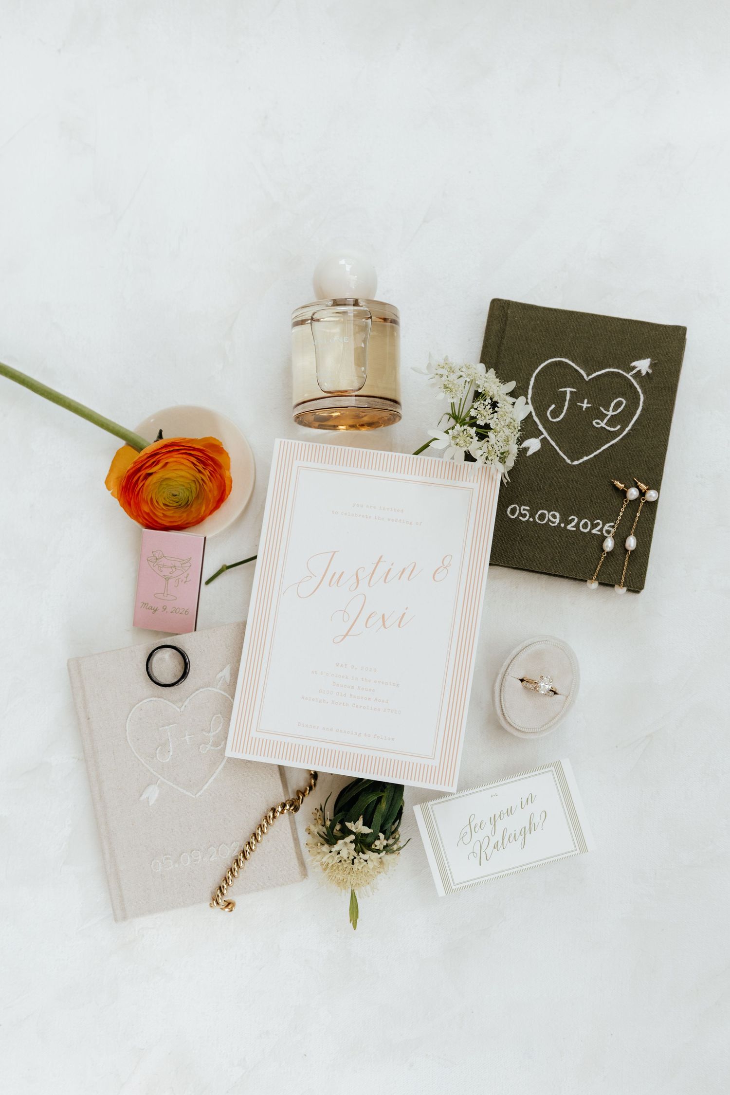 Elegant wedding flat lay with invitation, perfume, rings, floral accents, and monogrammed vow books dated 05.09.2026.
