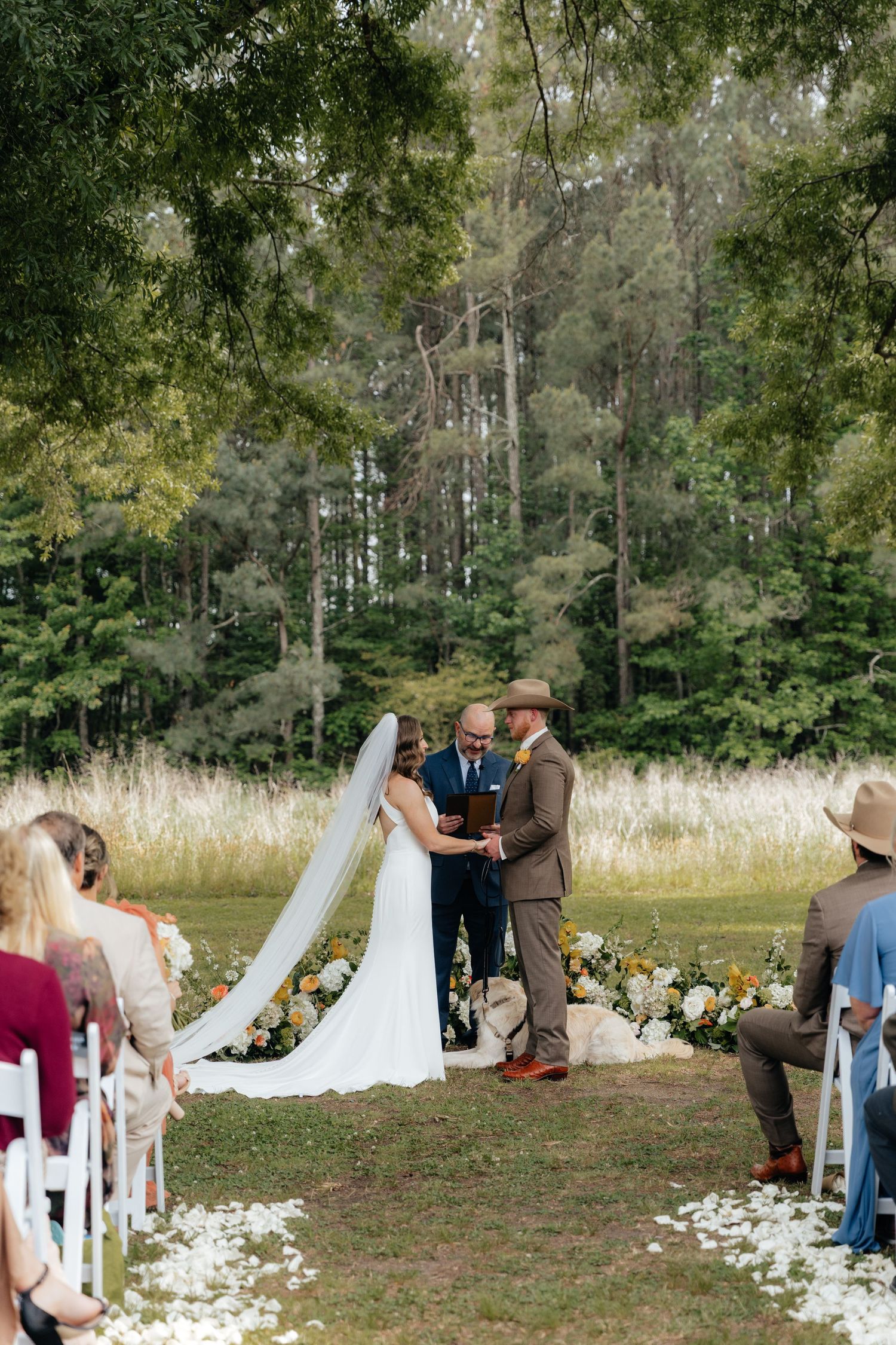 Outdoor wedding ceremony in a forest clearing with bride and groom at floral altar, white petal aisle, and seated guests.