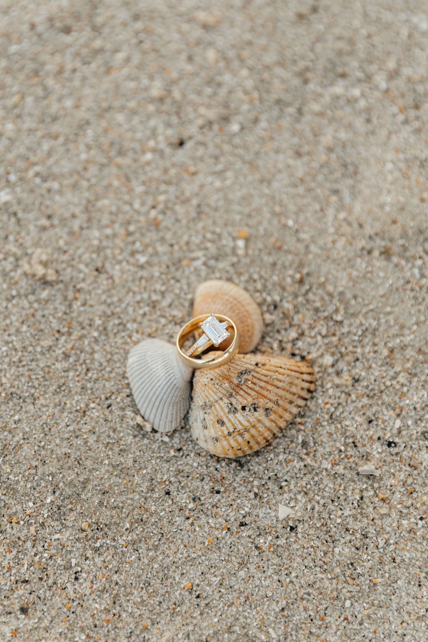 Gold diamond engagement ring resting on seashells in sandy beach setting.