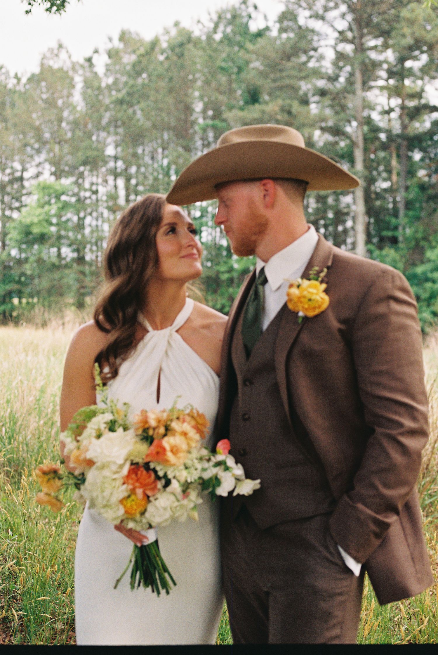 Bride and groom in western attire share a romantic moment outdoors, with a colorful bouquet and pine trees in background.