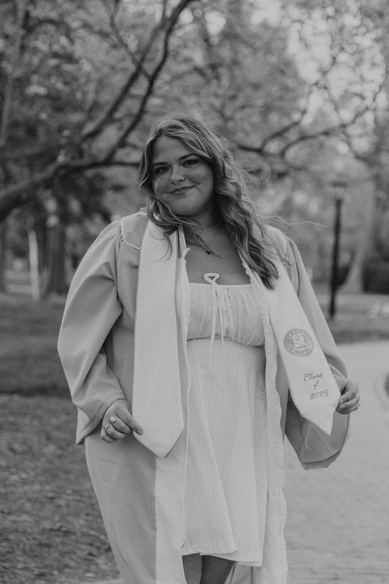 Black and white photo of a smiling graduate in cap and gown outdoors, holding her diploma, Class of 2023.
