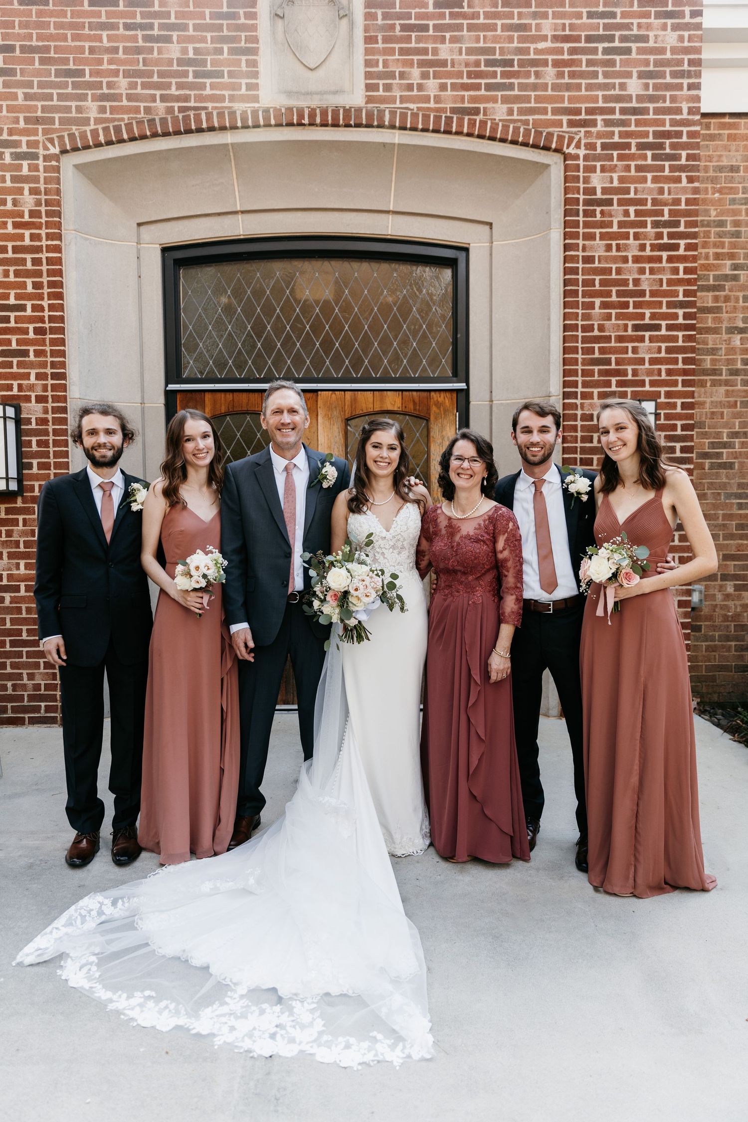 Wedding party in burgundy dresses and black suits pose in front of brick church exterior.