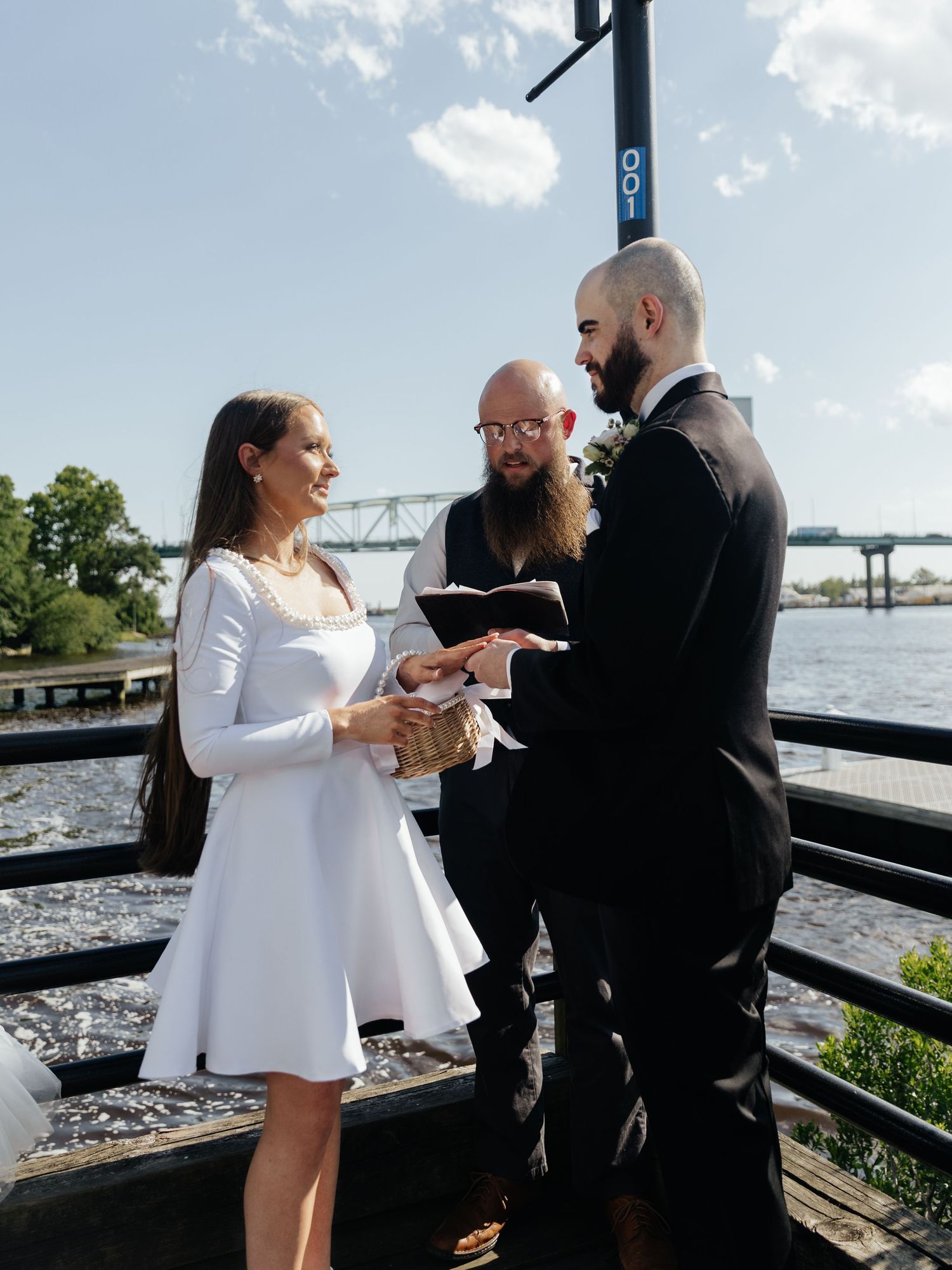 A wedding ceremony takes place on a wooden deck overlooking a river with a bridge in the distance.