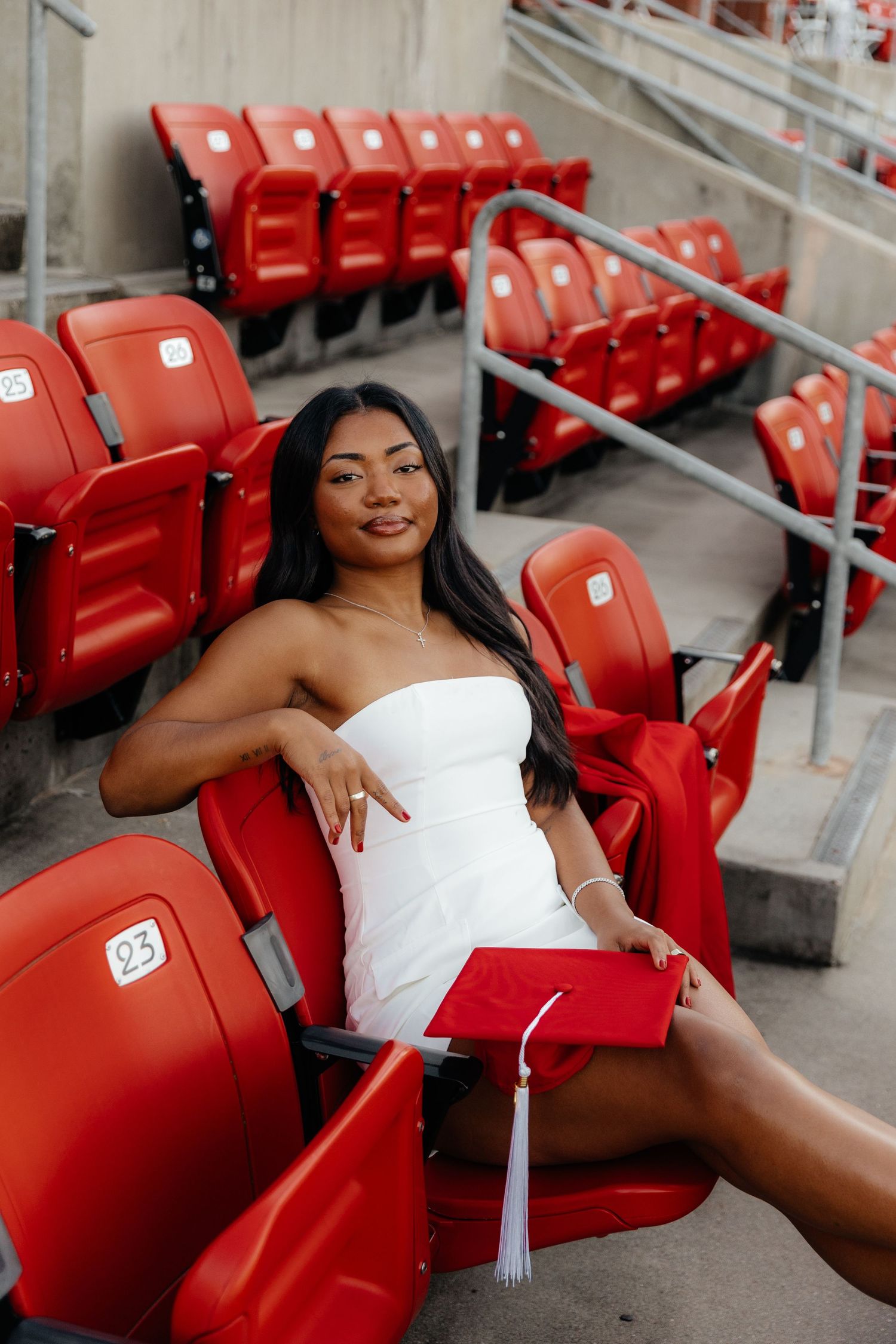 A graduate in a white dress poses on red stadium seats during a photo session.