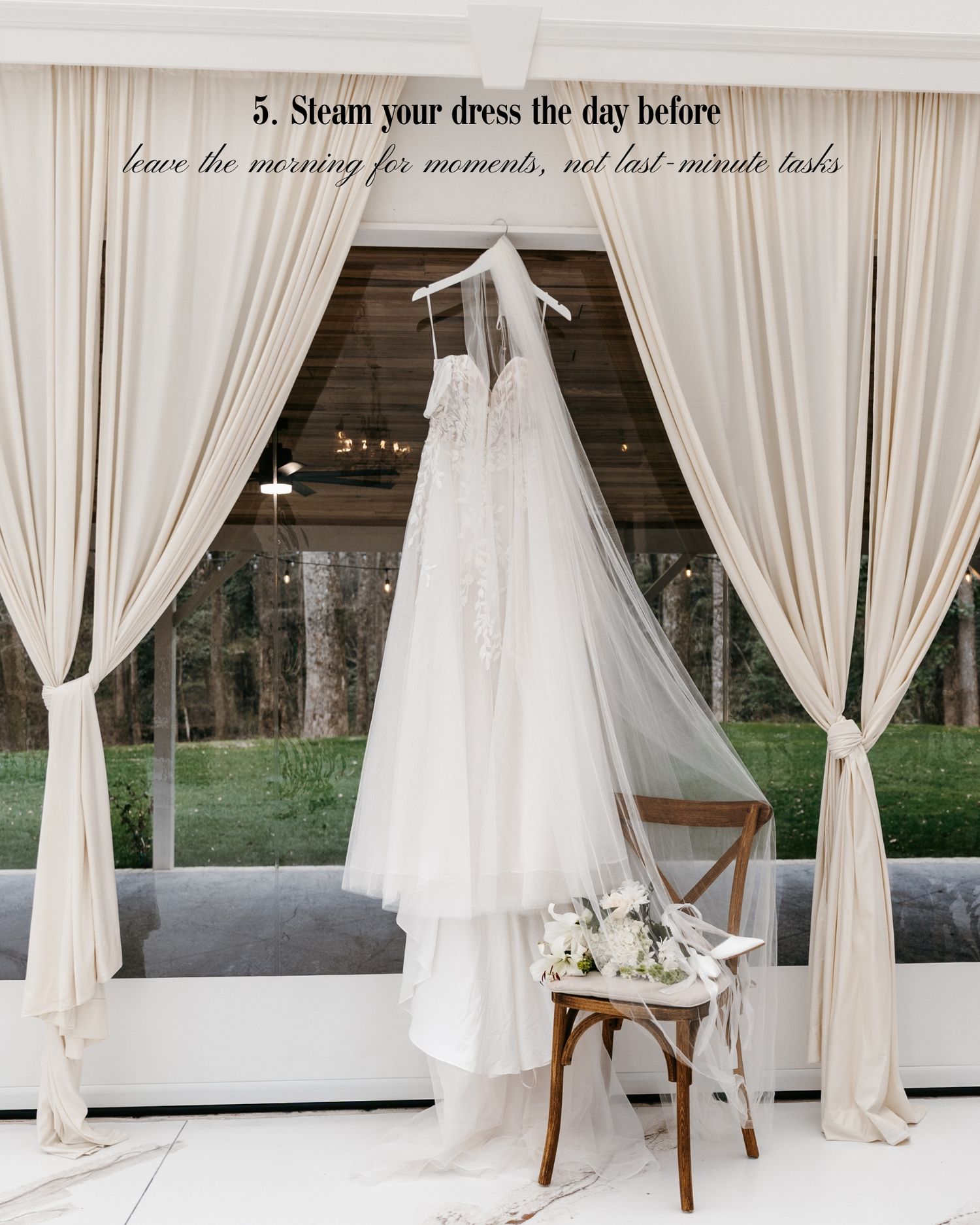 A lace wedding dress with veil hangs elegantly near draped curtains, with a floral bouquet on a wooden chair below.