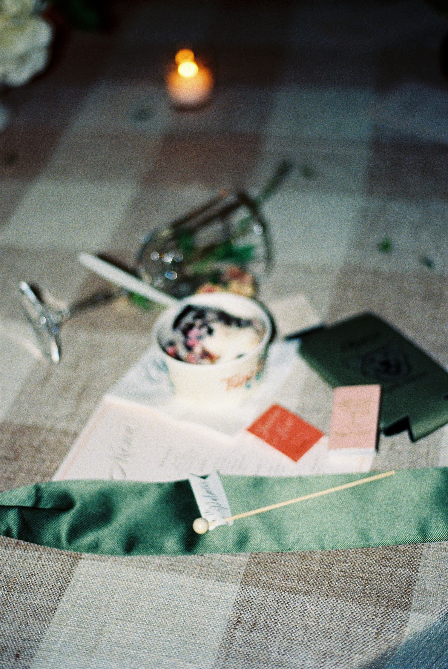 Elegant table setting with candle, floral cup, green ribbon, and stationery on plaid linen cloth.