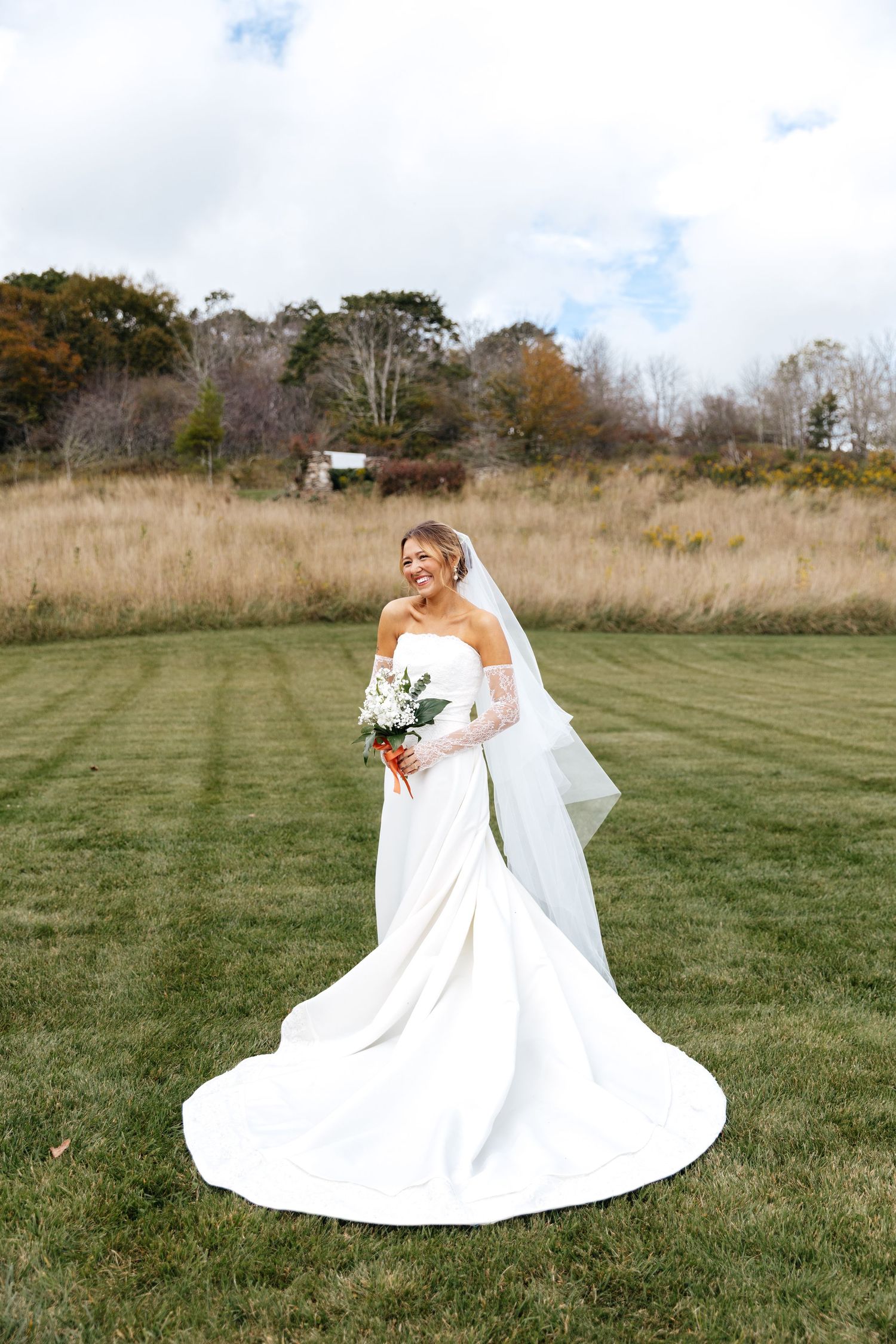 Smiling bride in a strapless white gown with long train and veil holds a bouquet on a green lawn.