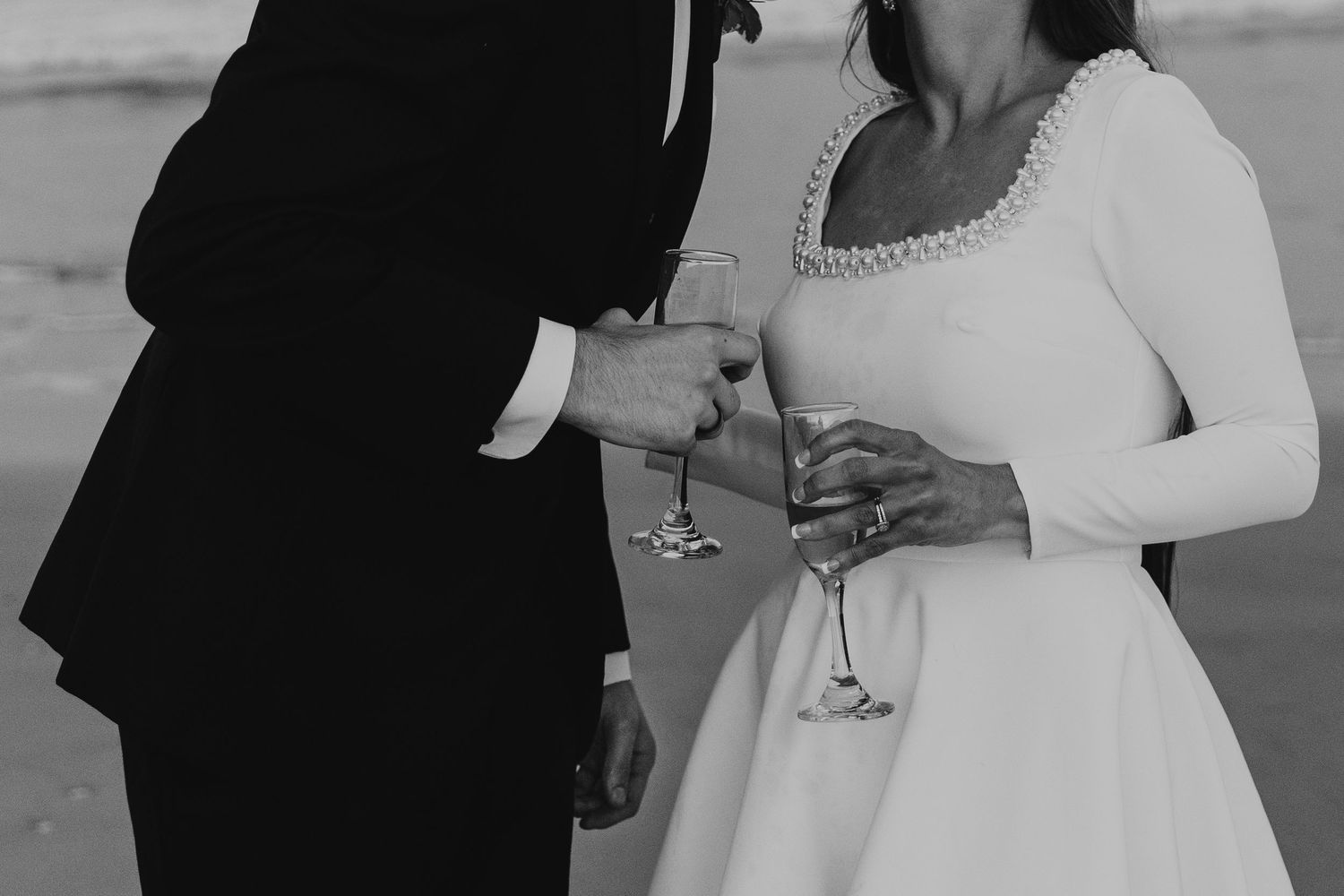 Black and white photo of a bride and groom in wedding attire clinking champagne glasses on a beach.