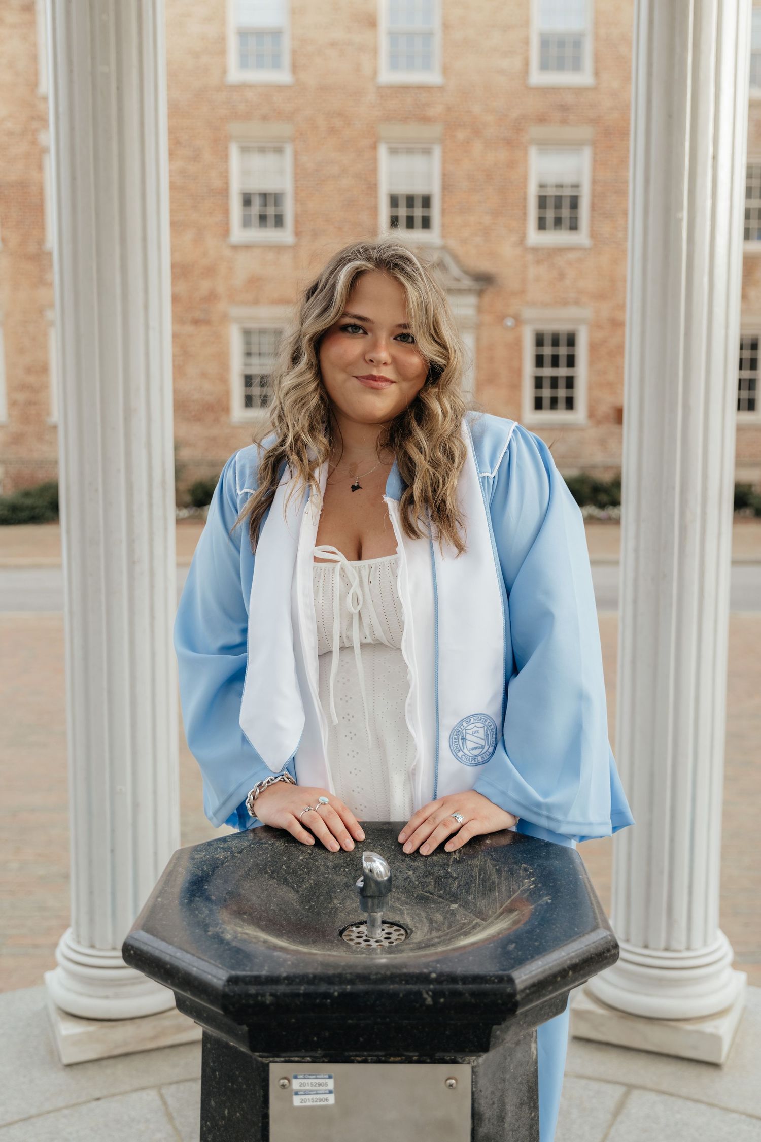 Young woman in light blue graduation gown poses at outdoor water fountain with brick building backdrop.