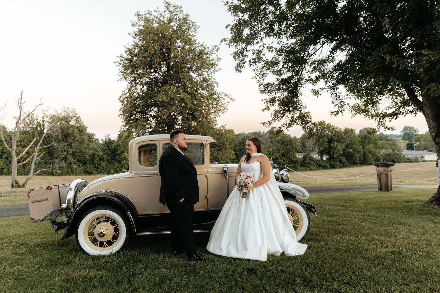 Wedding couple poses beside vintage cream-colored car on grassy lawn.