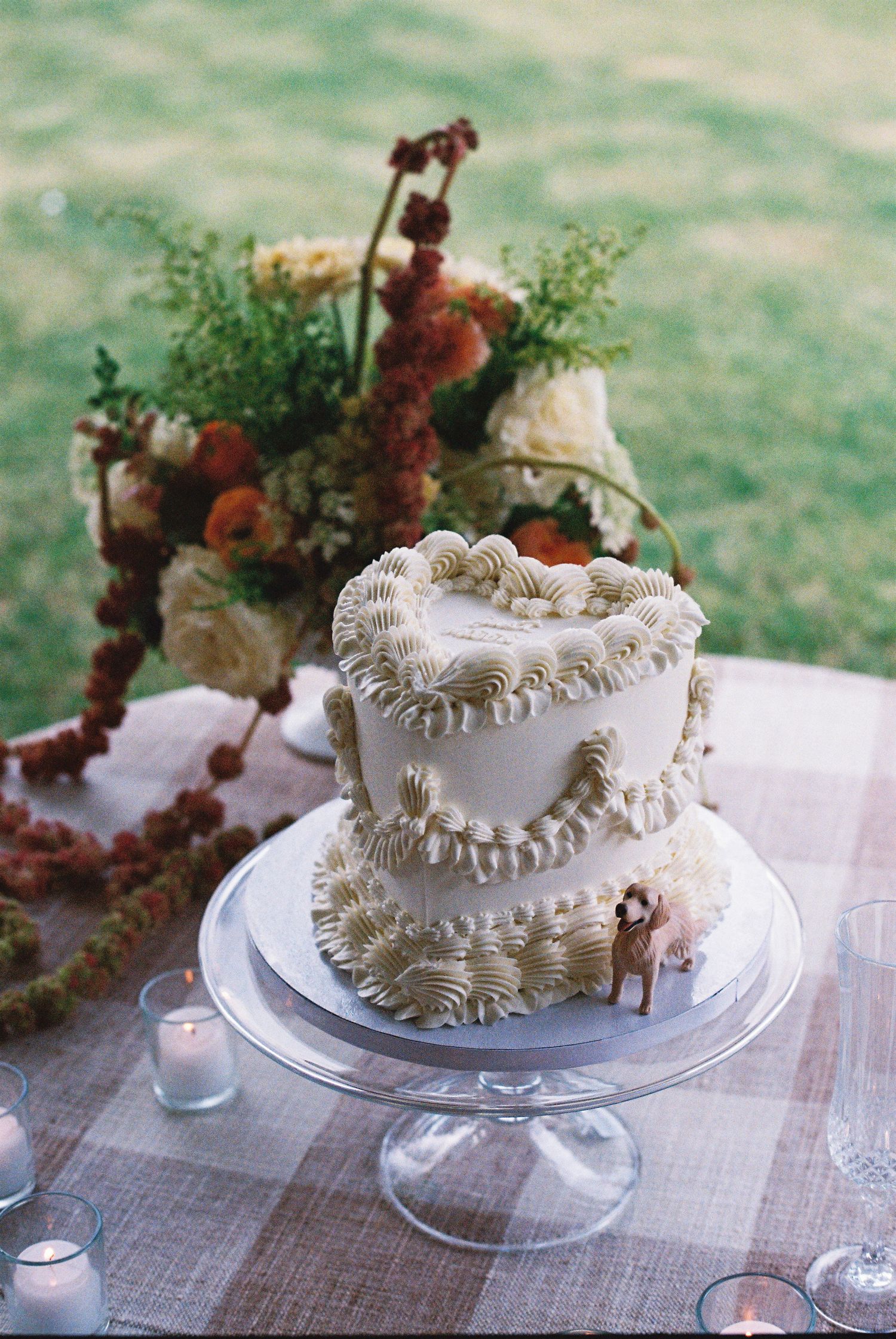 Two-tier white wedding cake with ruffled frosting and deer topper on glass stand, surrounded by autumn floral arrangements.
