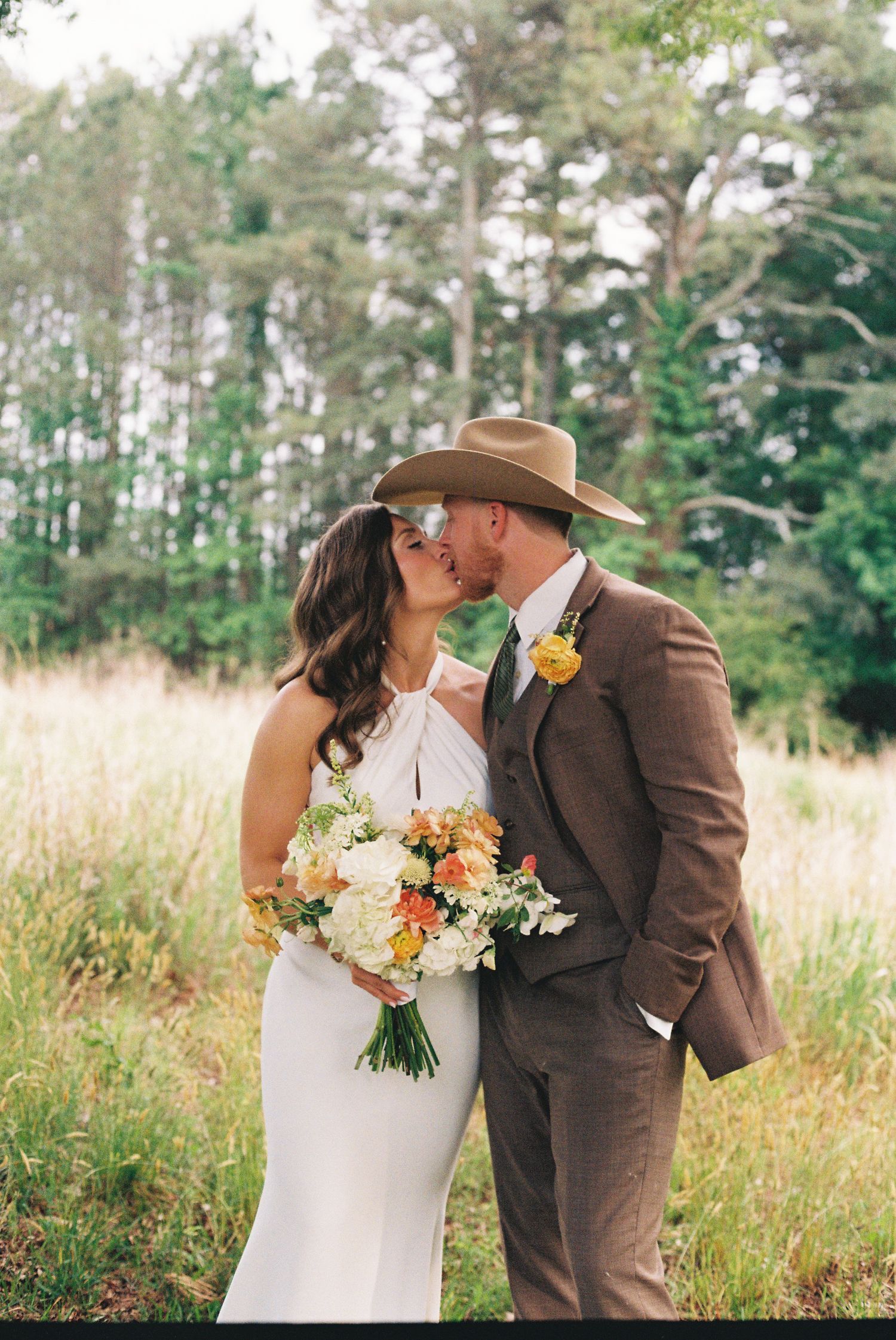 Bride and groom share a kiss in a sunlit meadow, she holds a colorful bouquet, he wears a brown suit and cowboy hat.