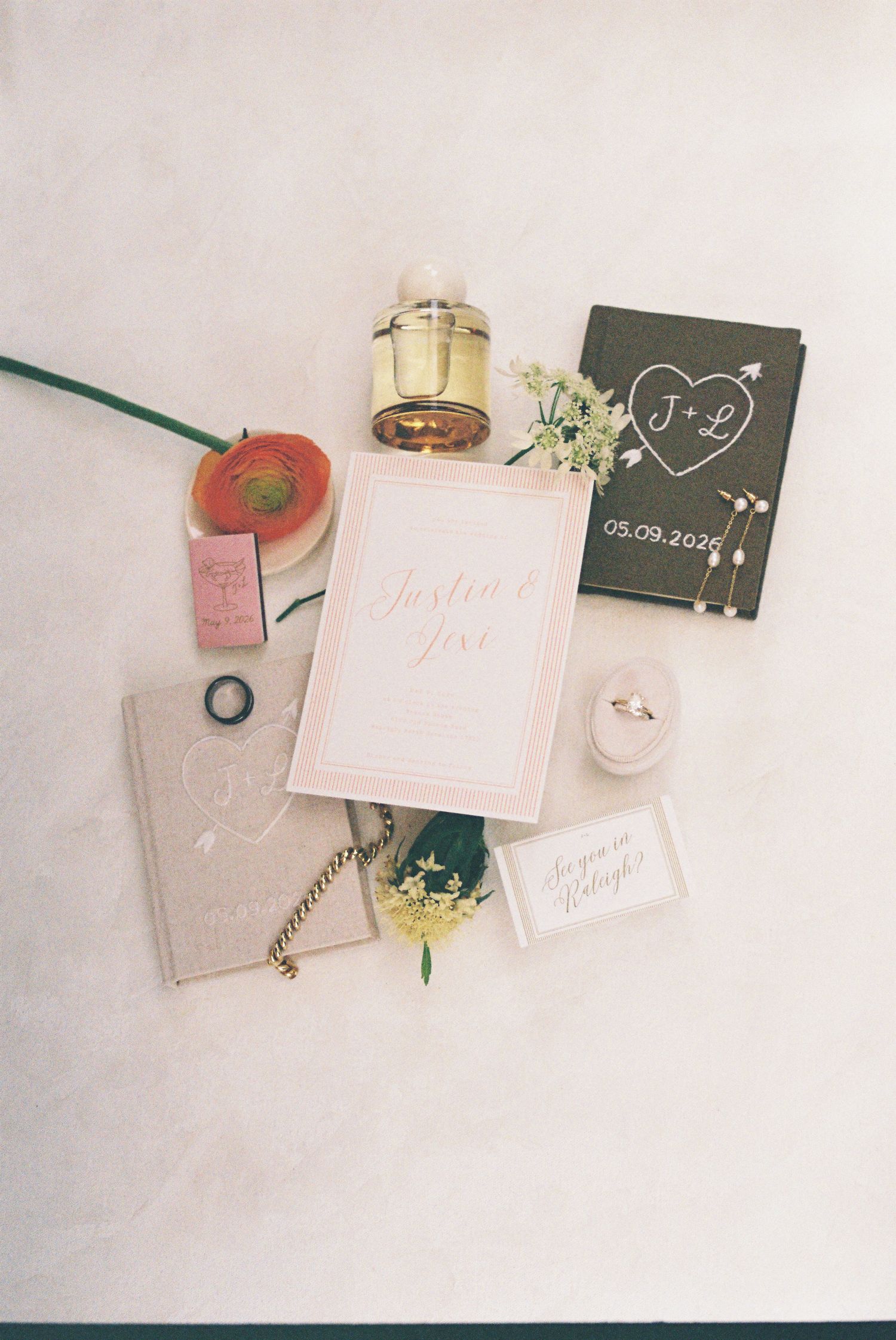 Elegant wedding flat lay with pink invitation, dark green monogram book, ring box, perfume, and floral accents on neutral background.