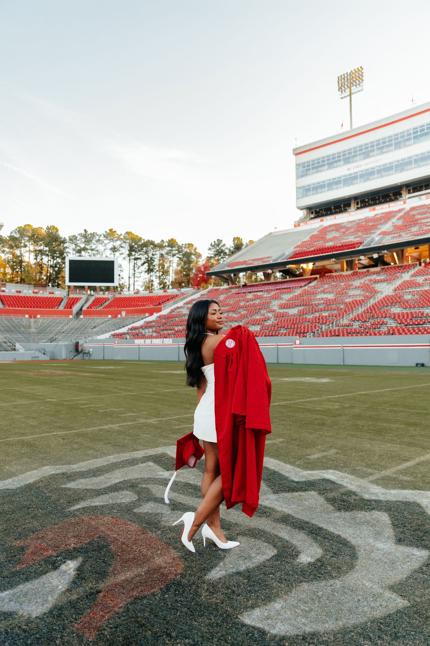 Young woman in white dress holding red graduation gown on football field stadium.