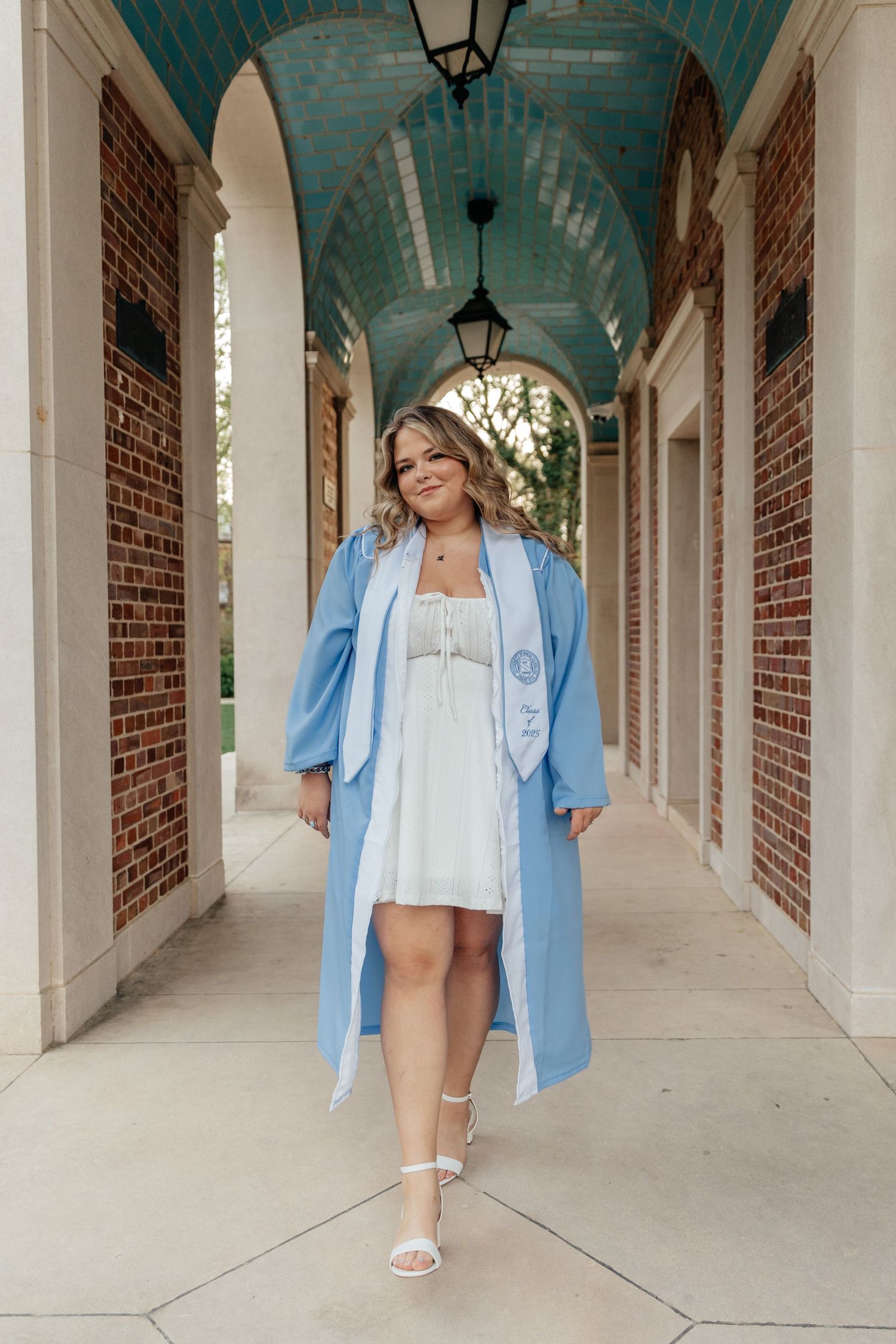 Smiling graduate in baby blue commencement robe and white dress posing in brick colonnade.