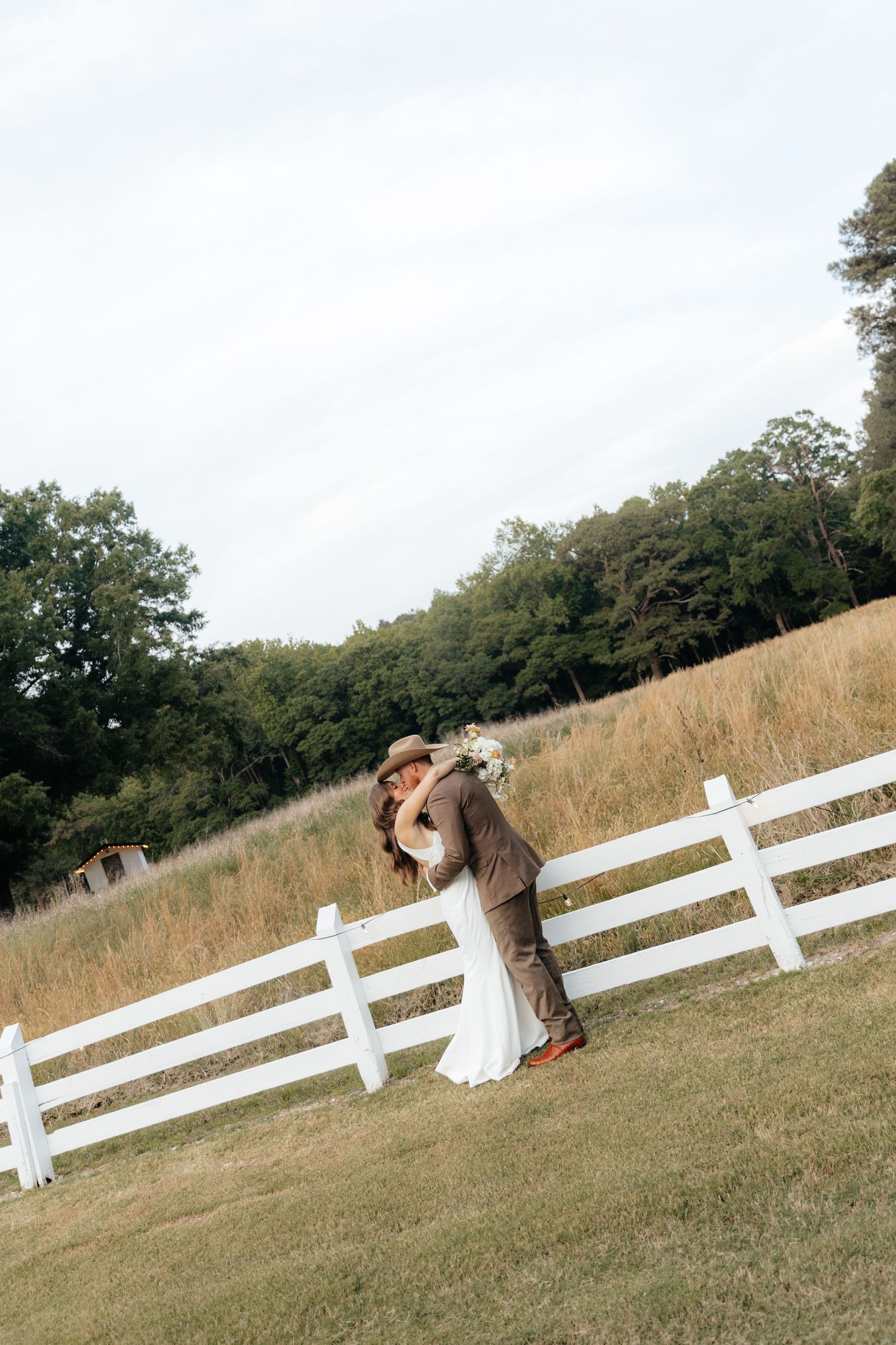 Bride and groom share a romantic kiss by a white fence in a rustic countryside setting on their wedding day.