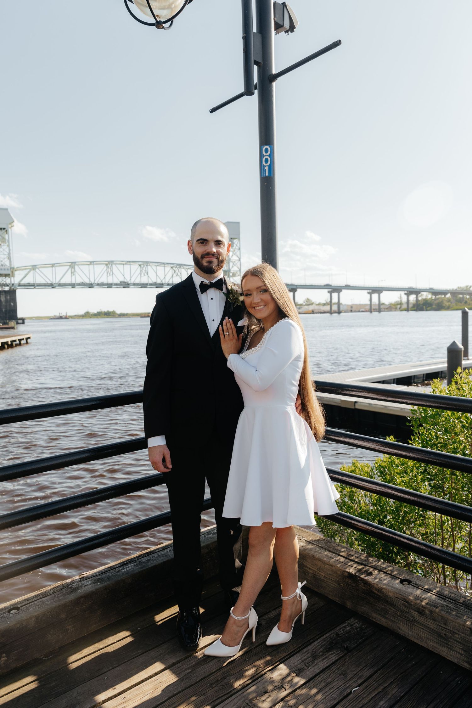 A newlywed couple poses on a wooden deck with a bridge and waterway in the background.