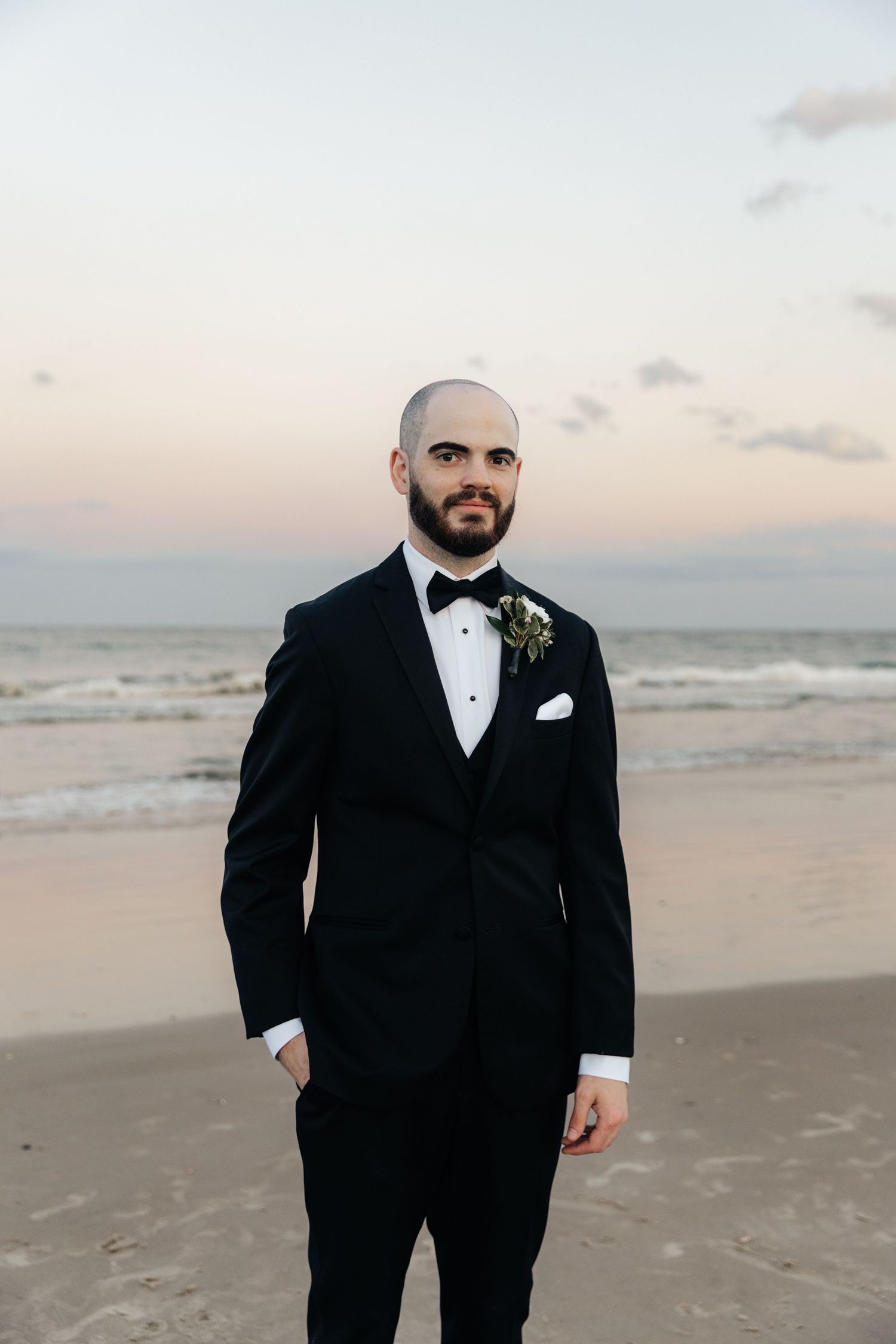 A groom in a classic black tuxedo poses on the beach at sunset.