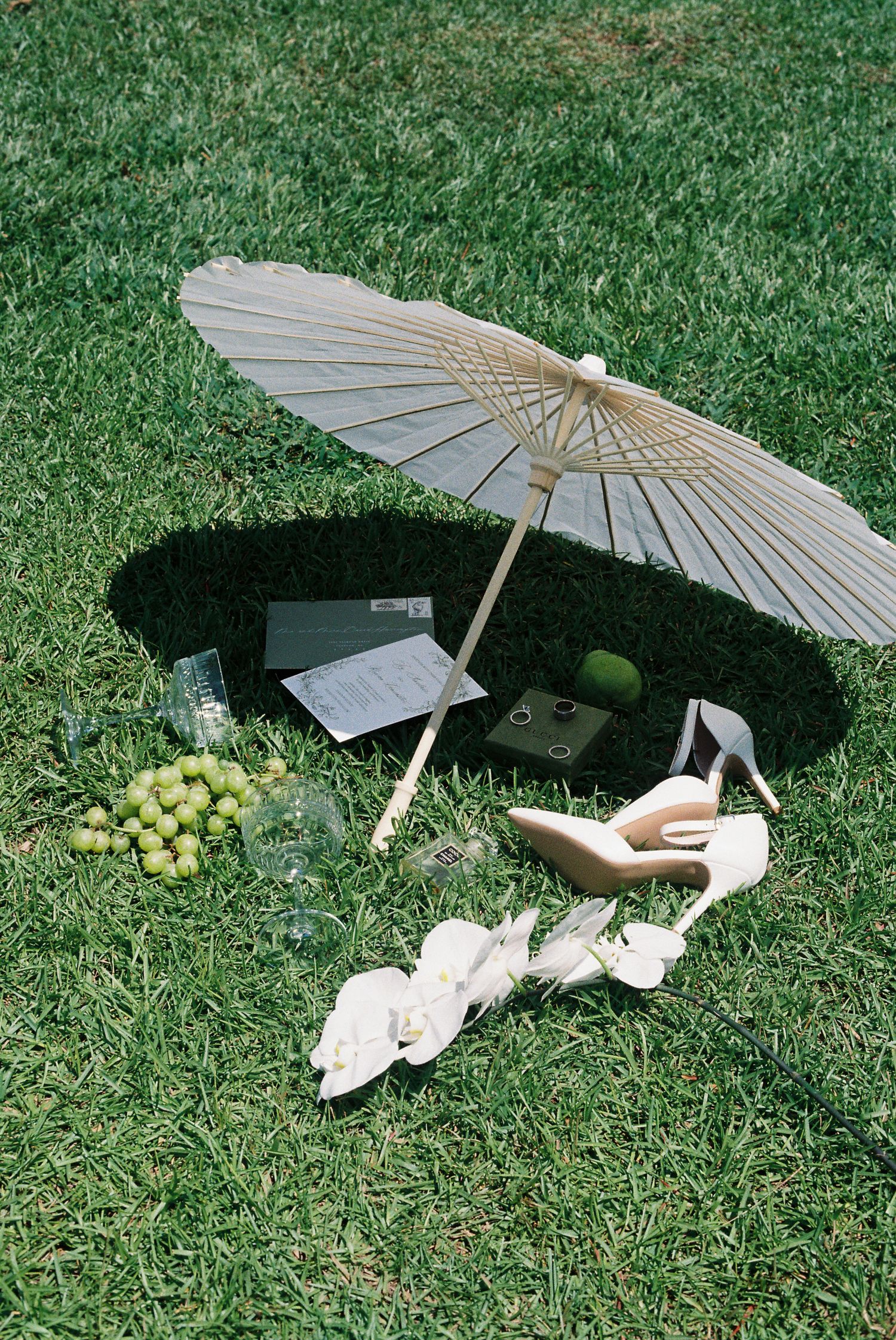White parasol, wedding stationery, and green grapes arranged artistically on grass.