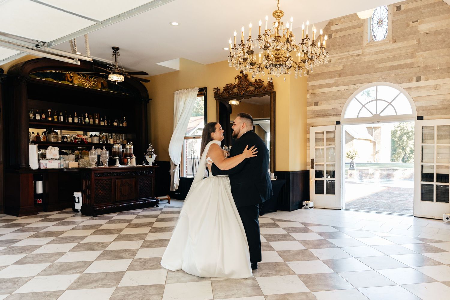 Wedding couple dancing on checkered marble floor under crystal chandeliers.