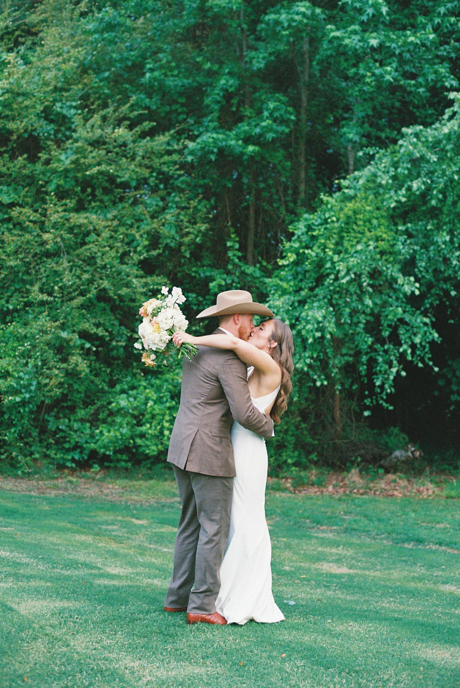 Bride and groom share a romantic kiss on a lush green lawn, with the bride holding a bouquet and groom wearing a cowboy hat.