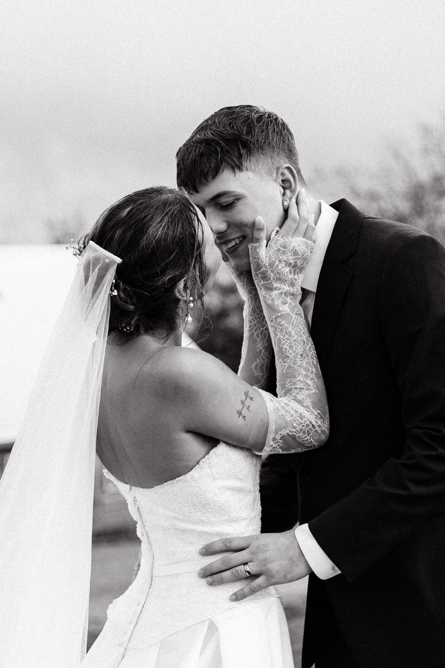 Black and white photo of a smiling groom in a suit and tattooed bride in a wedding dress sharing an intimate moment.