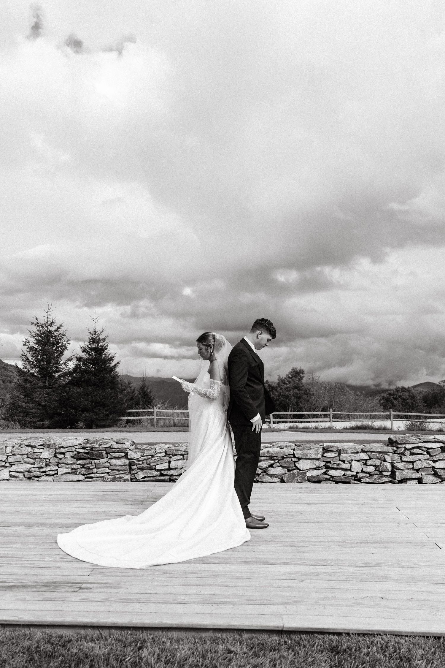 Black and white photo of bride and groom standing back-to-back on wooden deck with stone wall and cloudy sky backdrop.