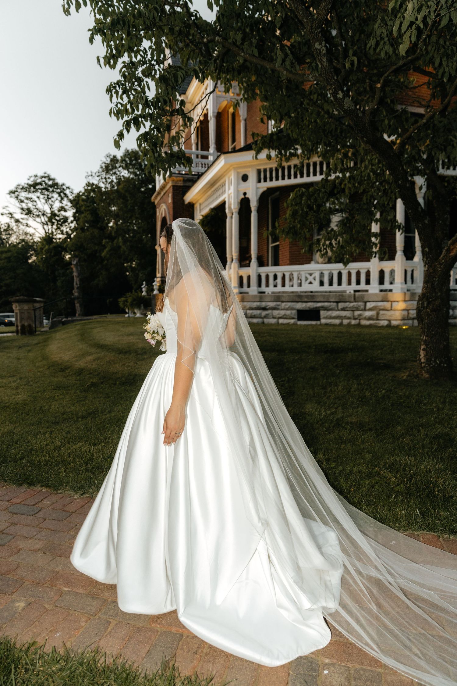 Elegant bride in flowing white gown and veil stands in front of historic Victorian mansion with wraparound porch.