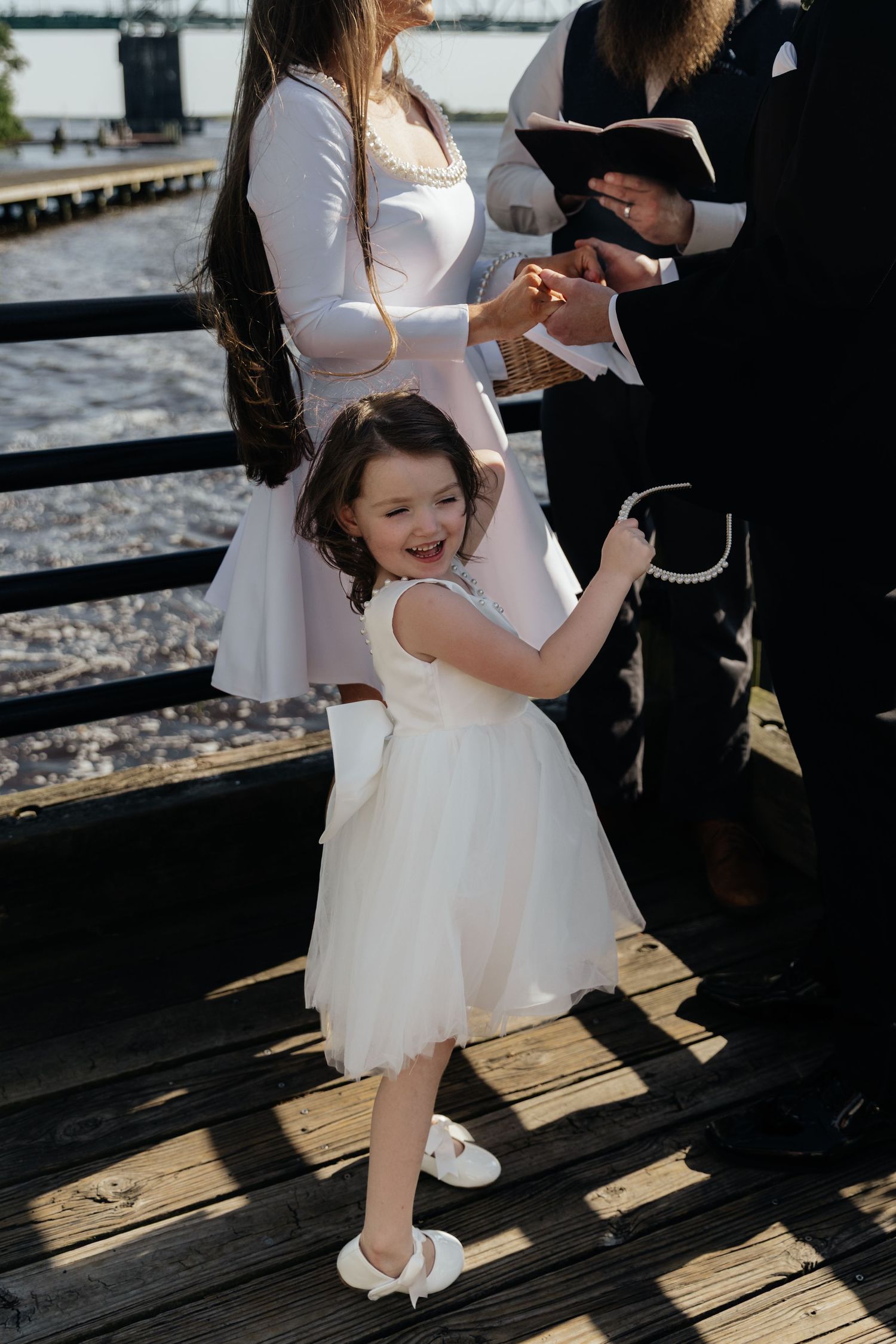 A young flower girl in white dress twirls during wedding ceremony by the water.