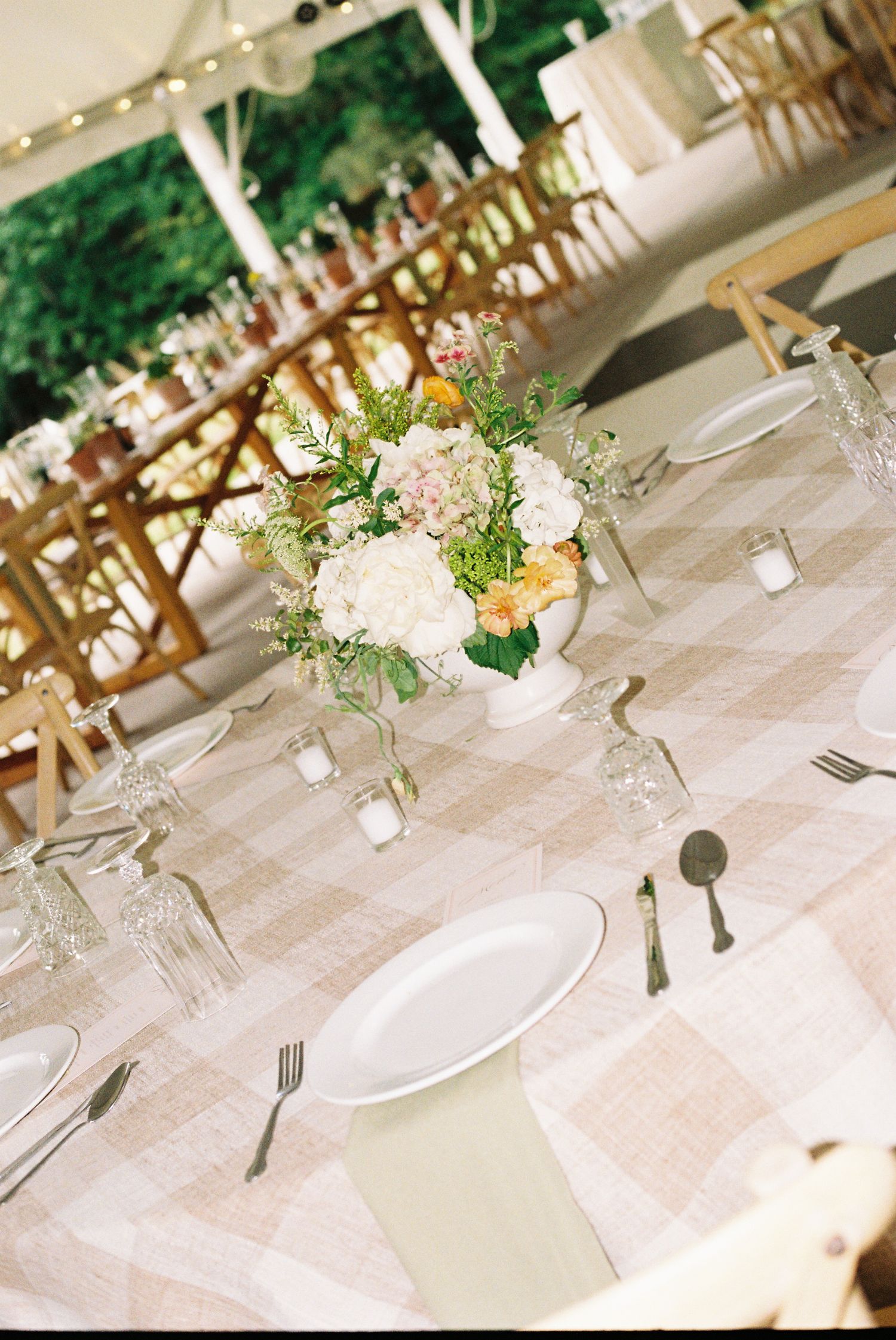 Elegant wedding reception table with pink gingham linen, white floral centerpiece, and glassware under a tent.
