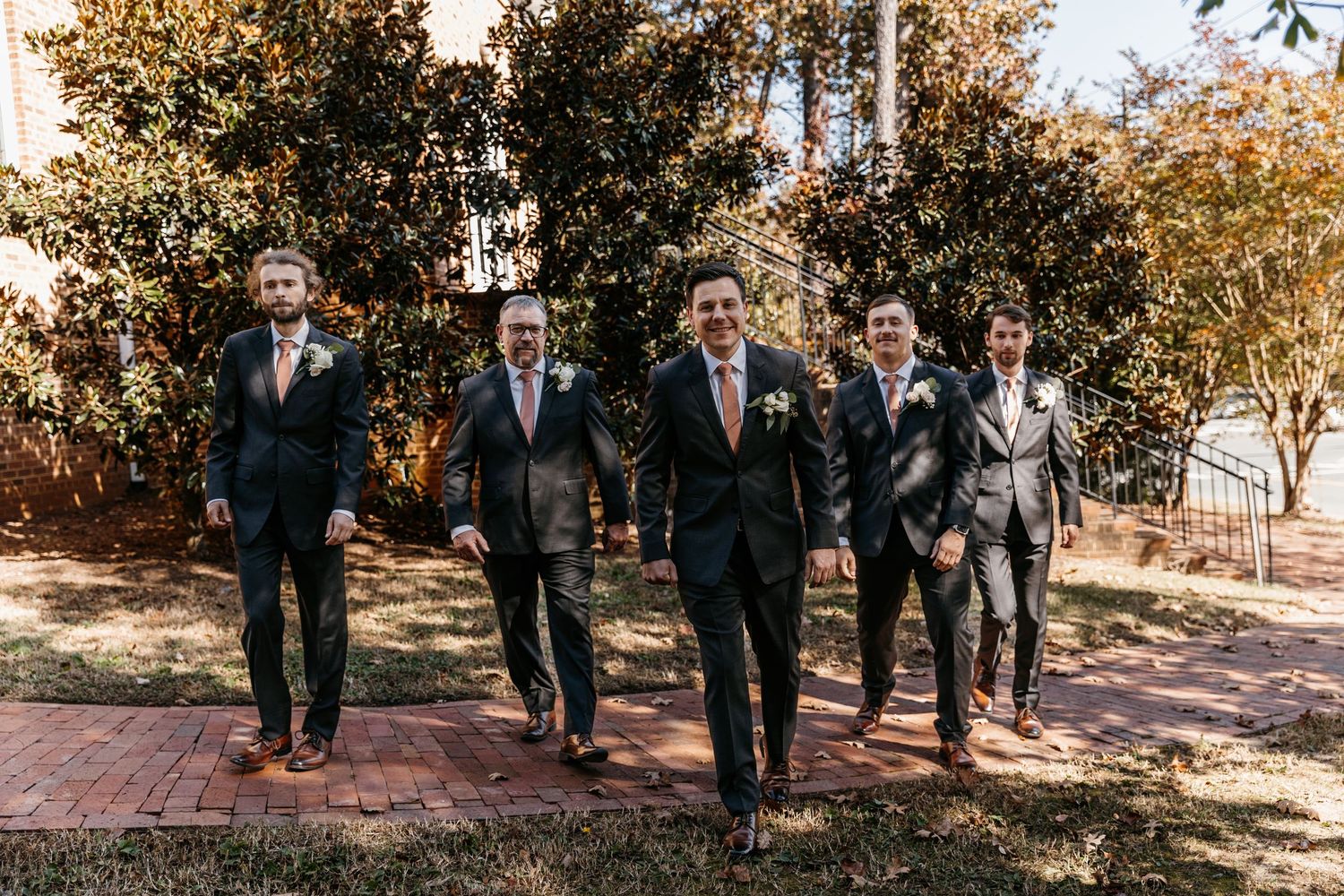 Groomsmen in dark suits with coral ties walking together on brick pathway.