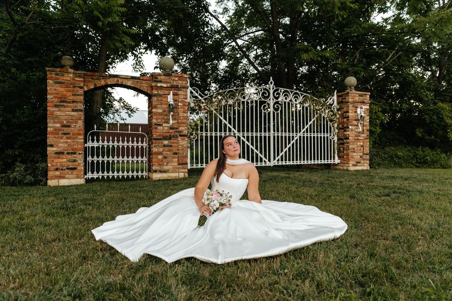 Wedding dress spreads beautifully across green grass near decorative iron gates.