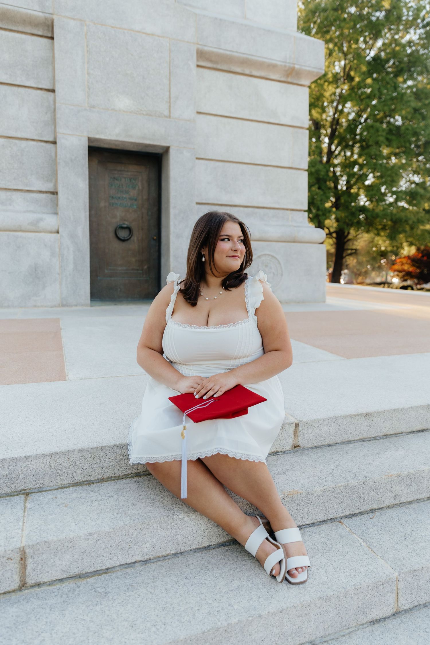 Graduate in white dress sitting on stone steps with red graduation cap.