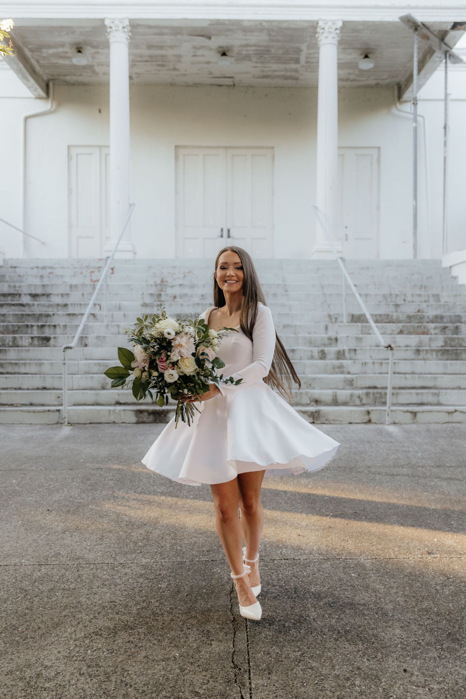 A bride in a short white dress twirls playfully in front of a white brick building.