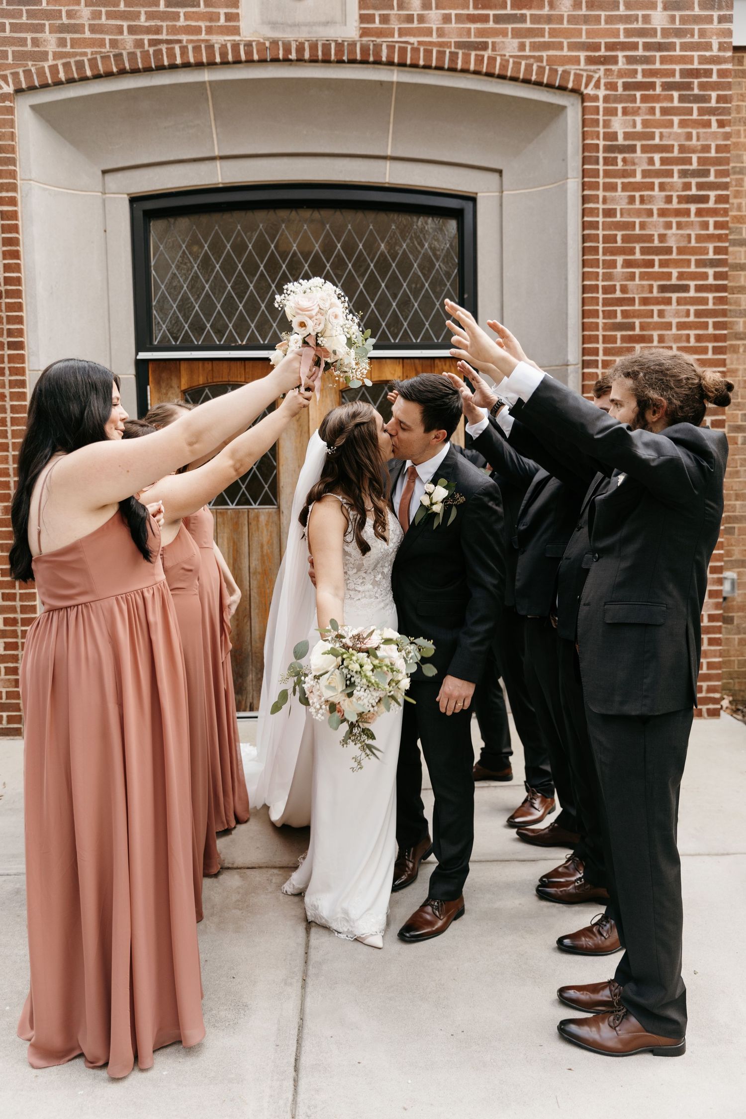 Wedding celebration with bridesmaids in coral dresses and groomsmen in black suits forming arch while the bride and groom walk through it.