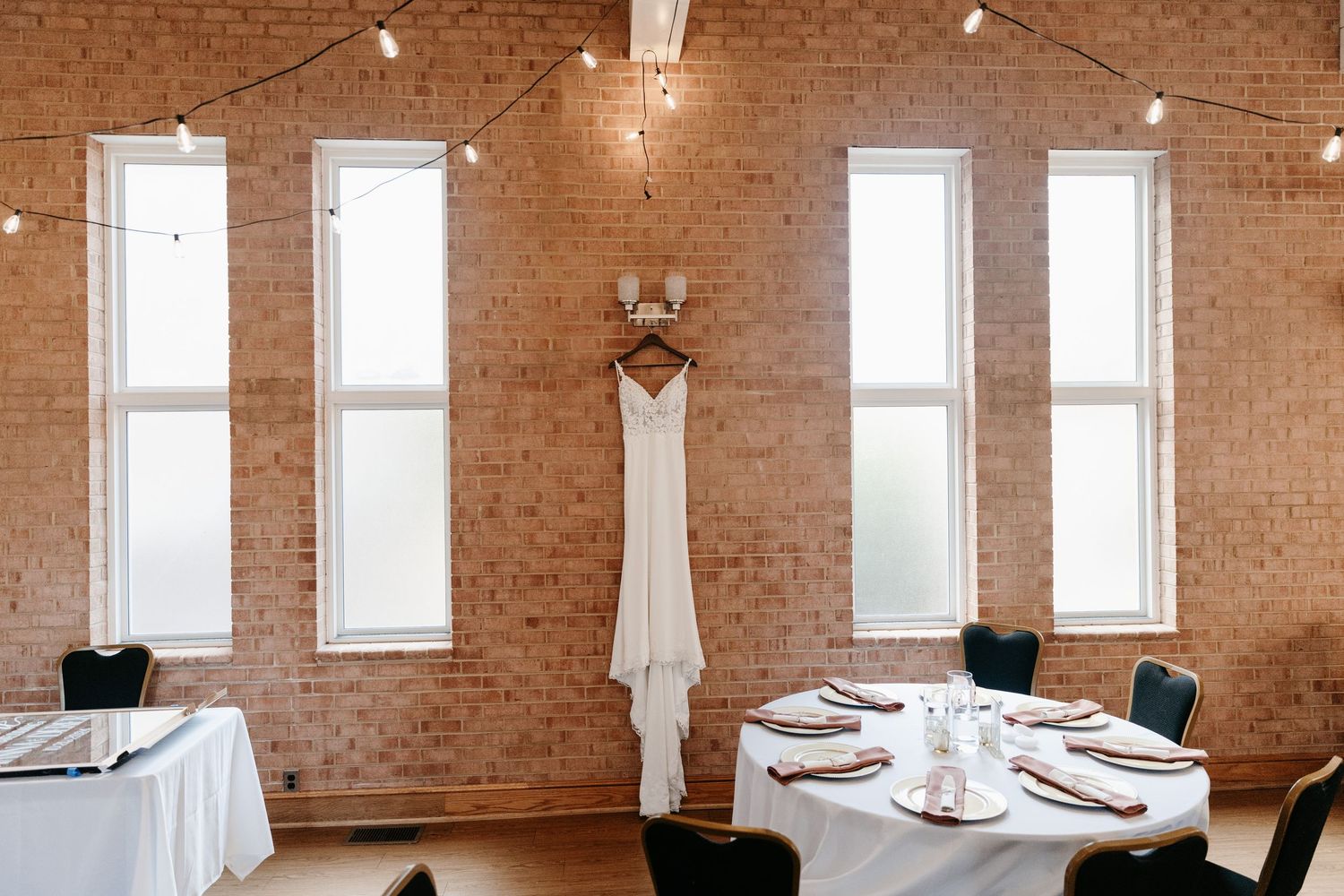 Wedding gown hanging inside a large industrial brick venue space with high ceilings and white windows for a wedding reception.