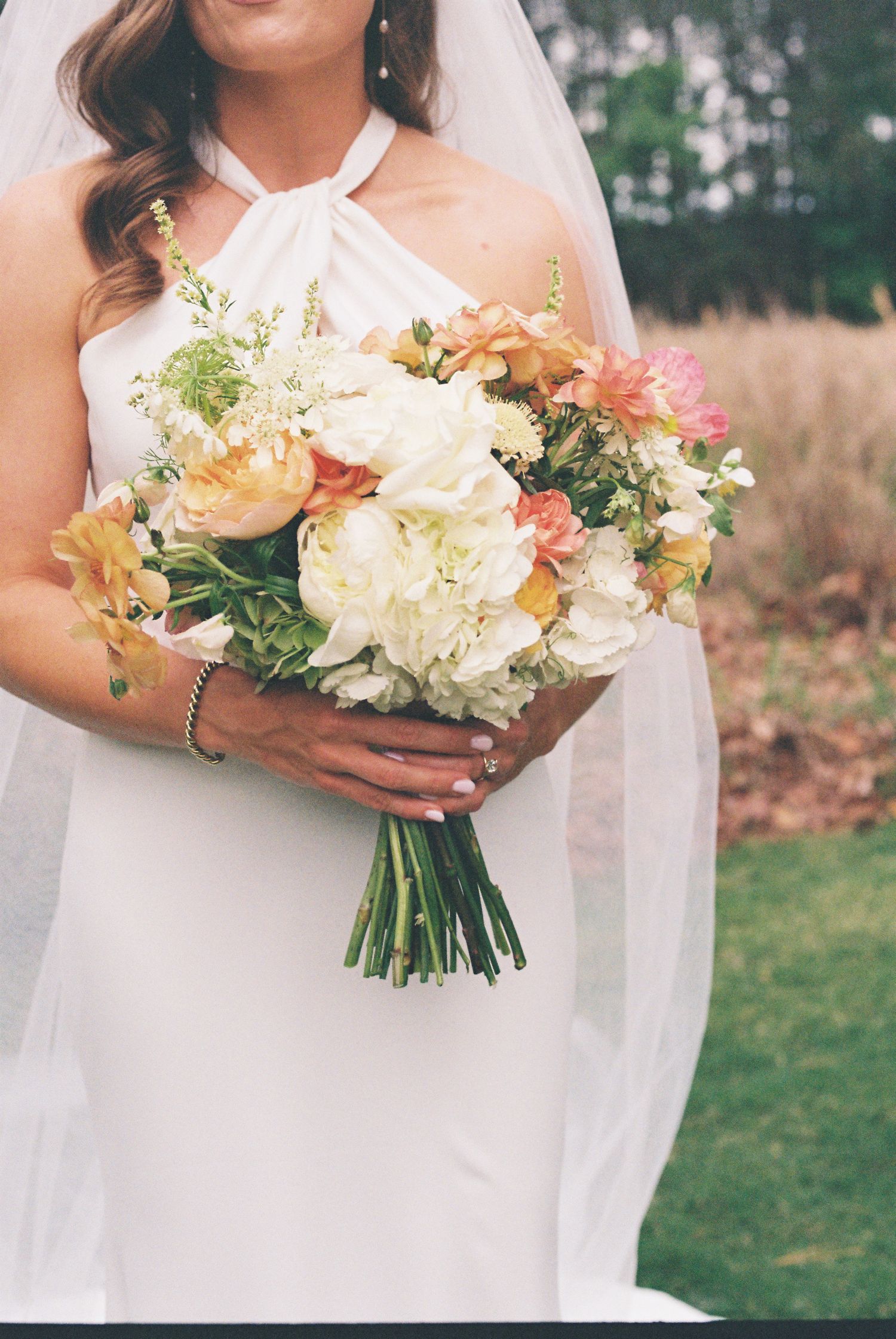 Bride in white halter gown holding lush bridal bouquet of peach, coral, and ivory flowers with greenery outdoors.
